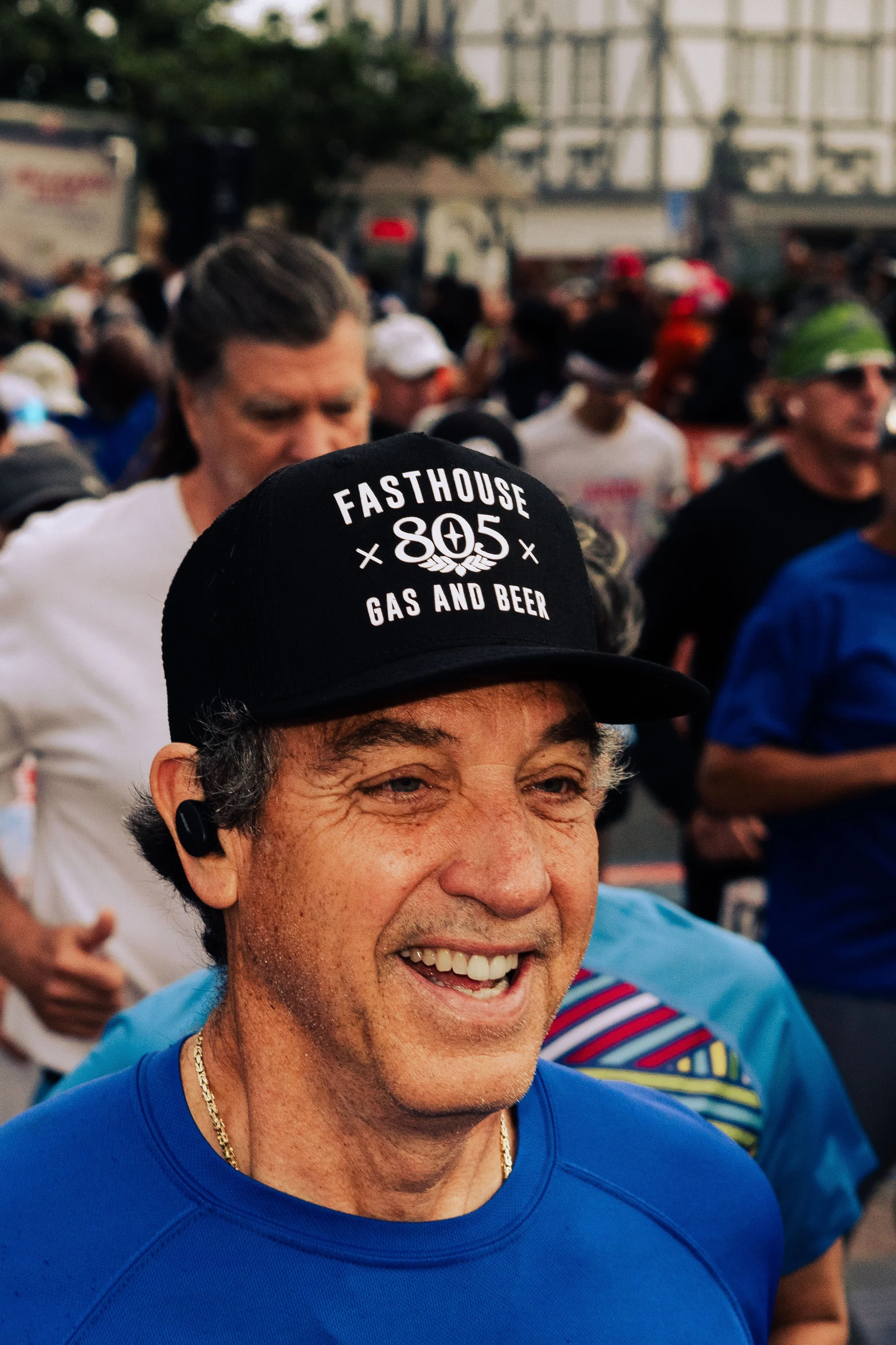 Smiling man wearing a blue shirt, gold chain, and a black cap with the text "Fasthouse 805 Gas and Beer" on it, running in a crowded outdoor event.