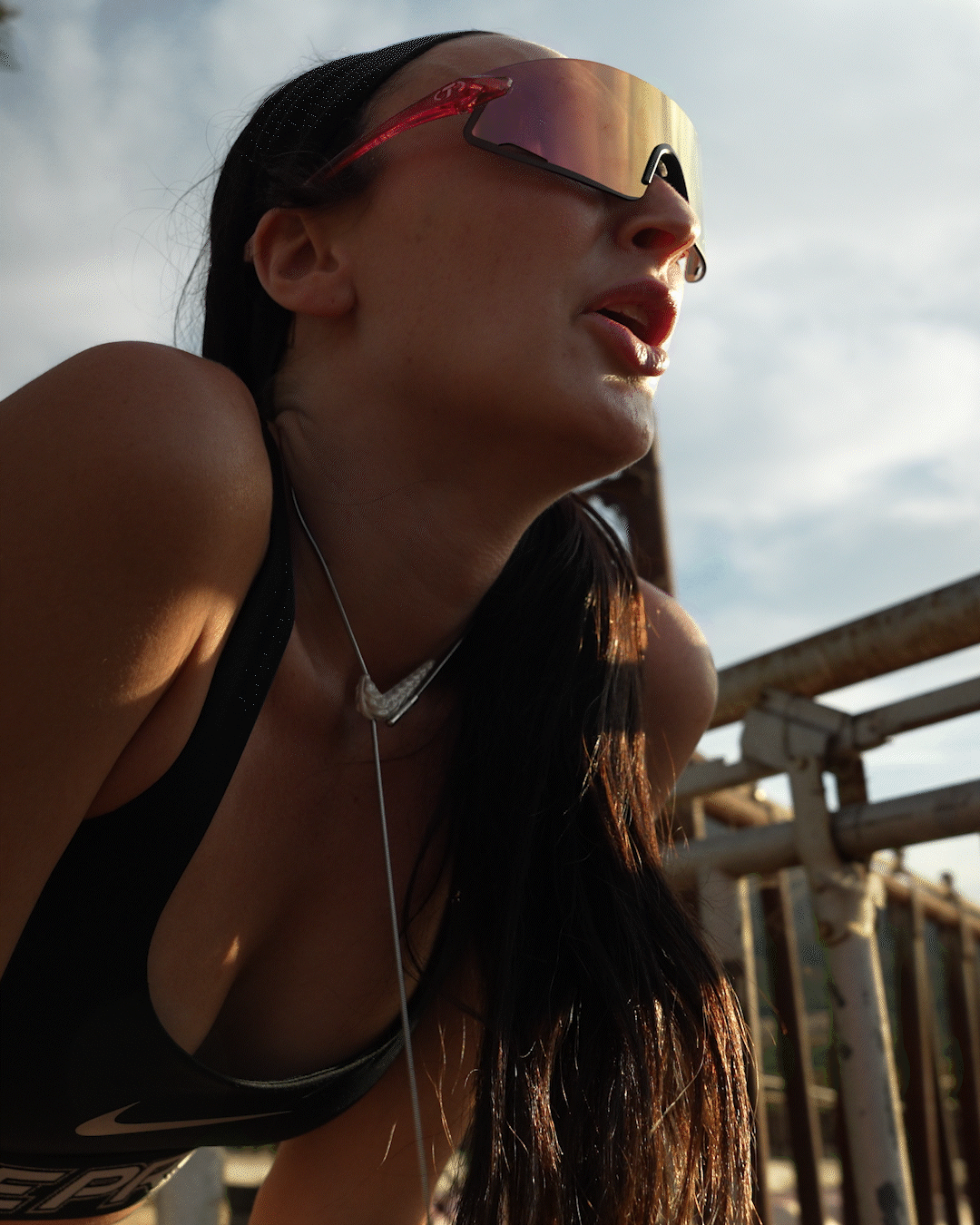 Close-up of a woman wearing sports sunglasses, a black athletic top with the Nike logo, and earbuds, leaning on a rusty metal railing outdoors during sunset or late afternoon.