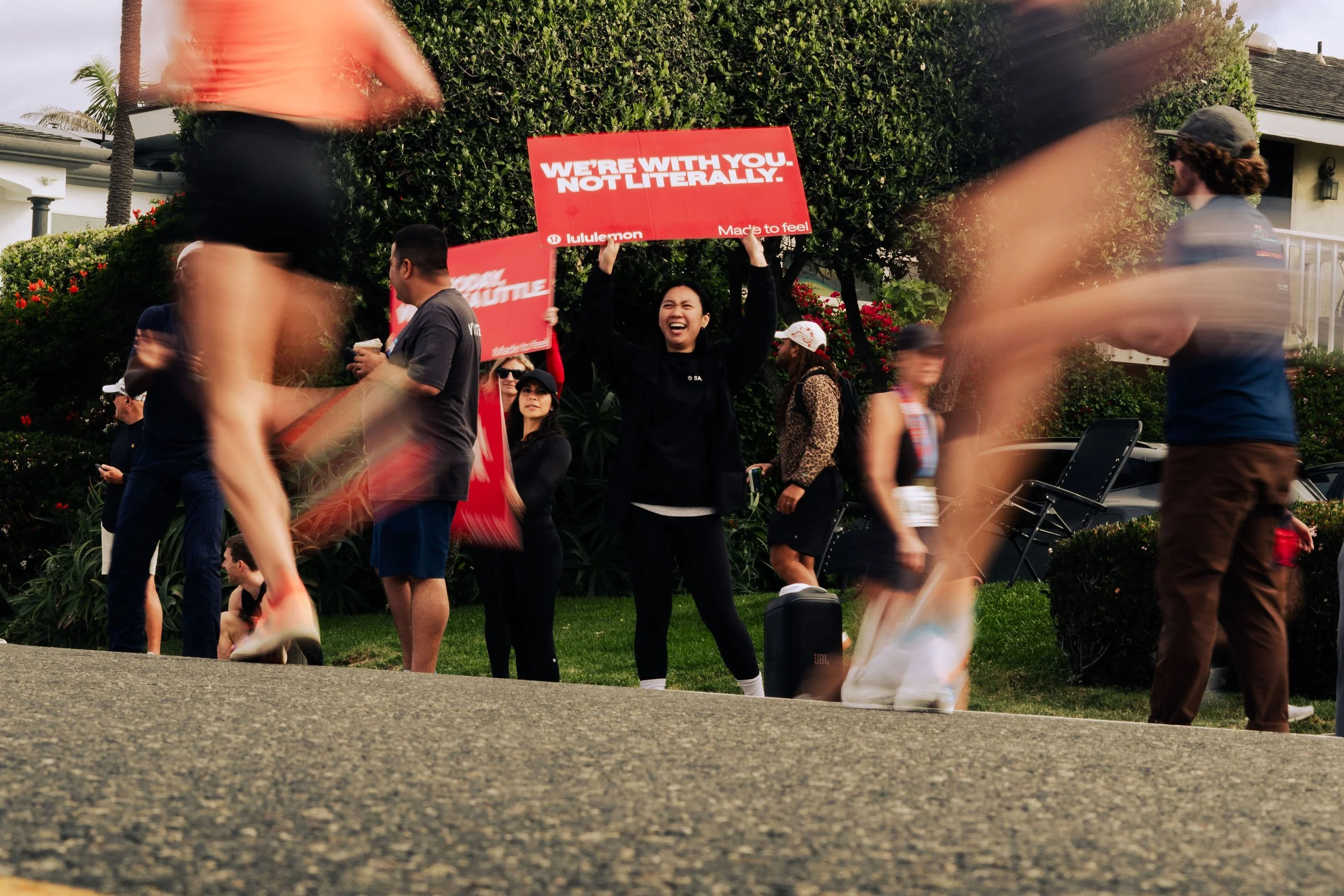 People participating in a relay race, with one woman smiling and holding a red sign that says 'WE'RE WITH YOU. NOT LITTERRALLY.' in a suburban neighborhood setting.