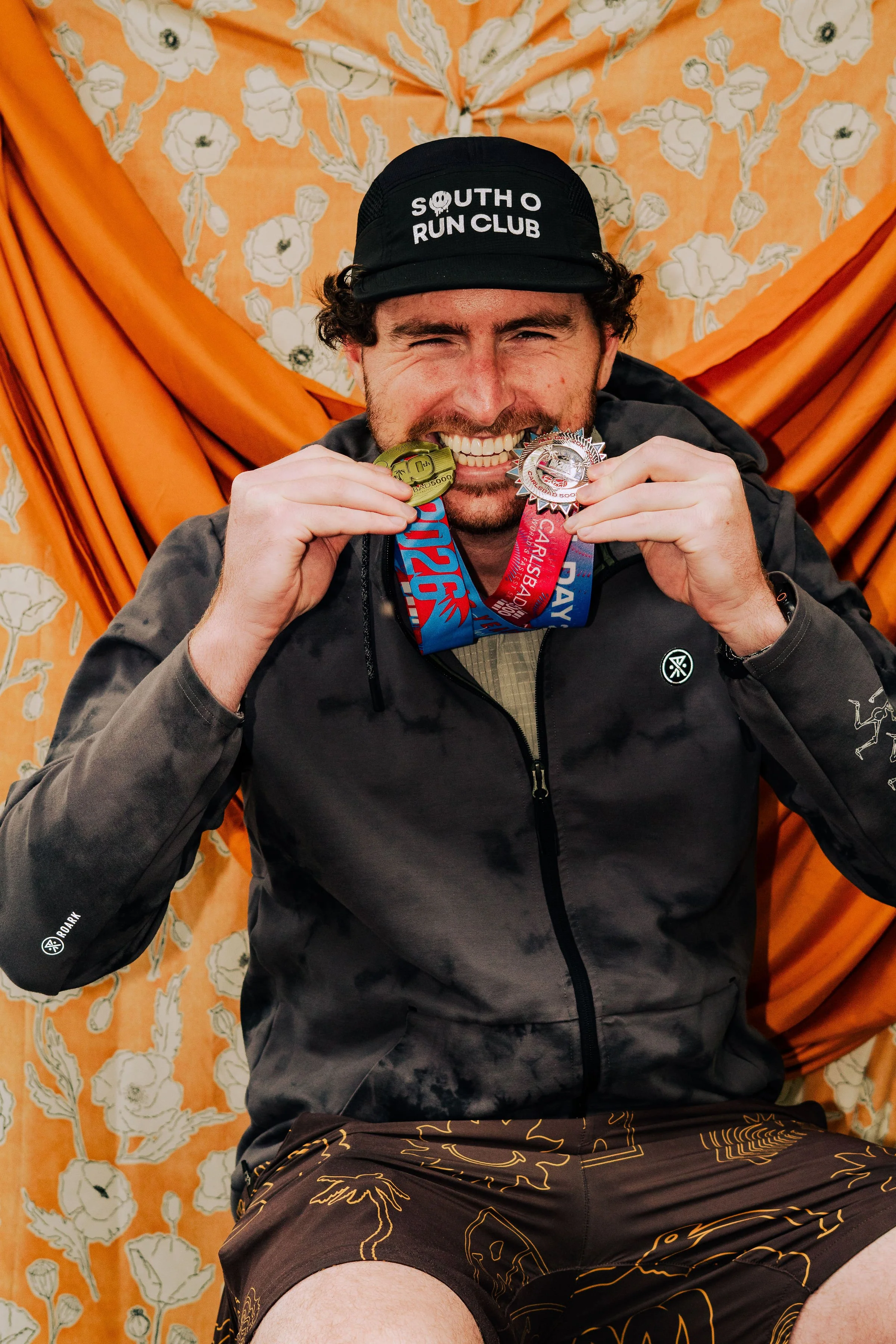 A man smiling and holding medals in his hands. He is wearing a black cap with white text that reads 'South O Run Club' and a black jacket. The background features orange fabric and a floral-patterned orange curtain.