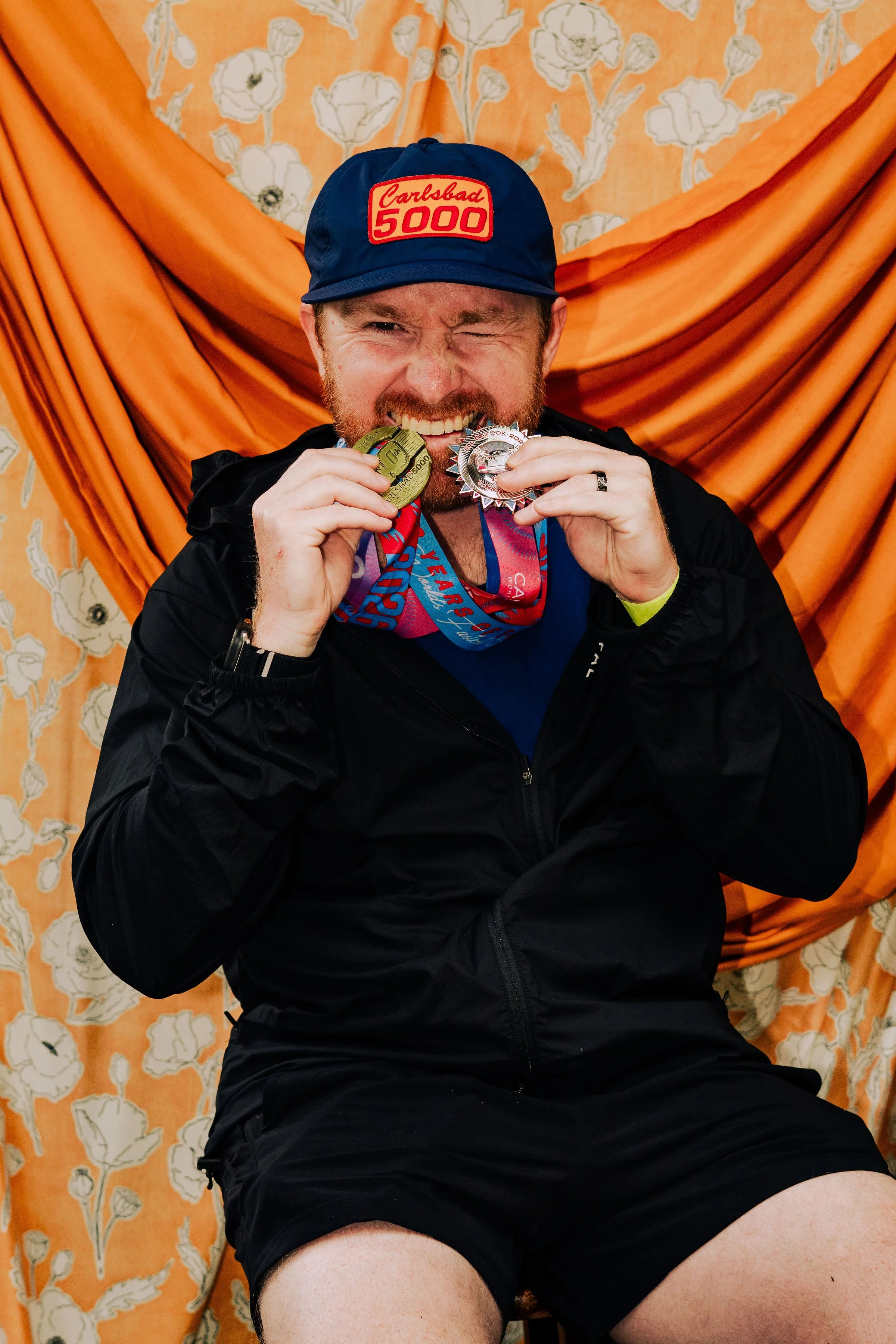 Man in a black jacket with medals around his neck, wearing a blue cap with red and yellow text, sitting against an orange and cream floral fabric backdrop, biting a medal.