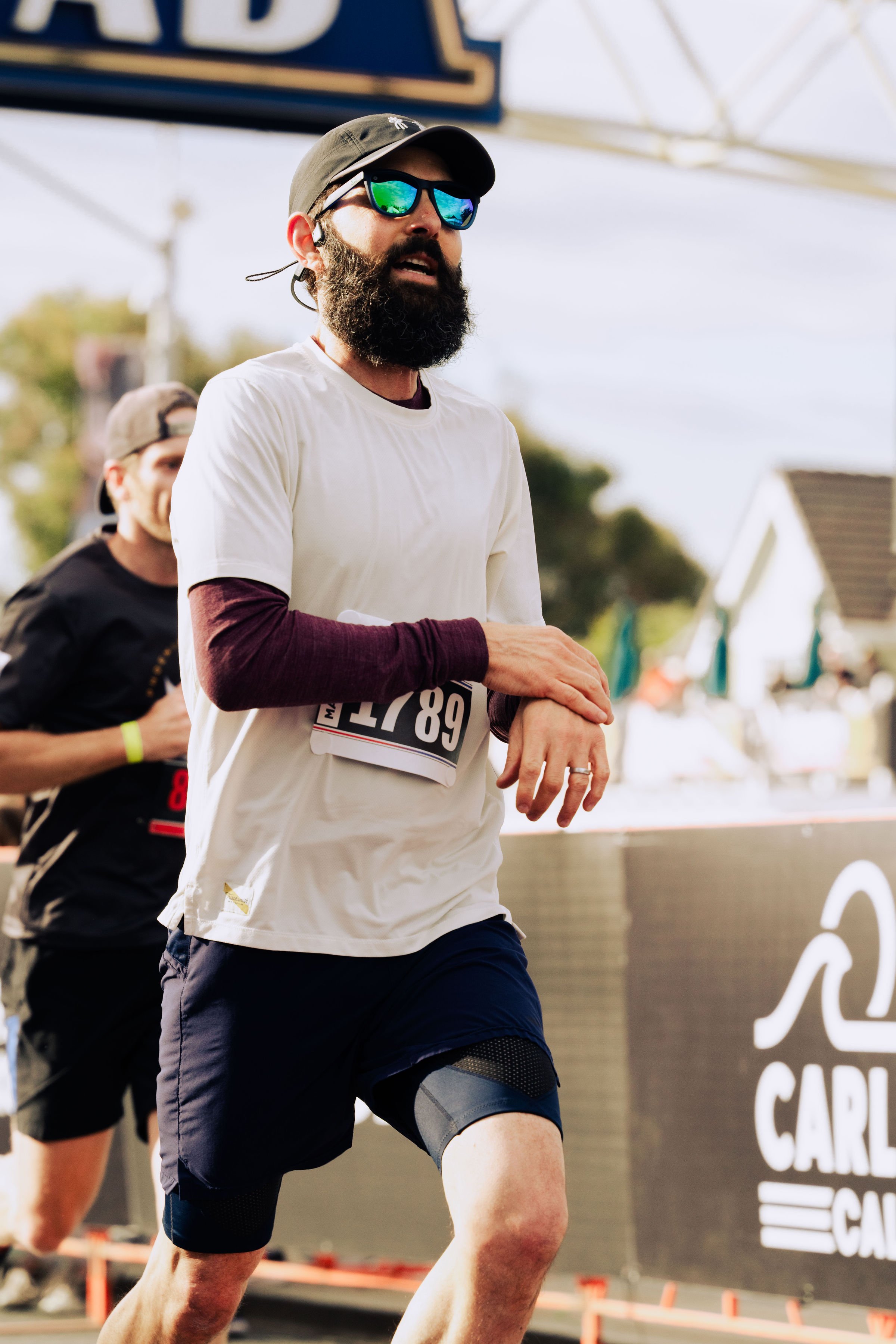 A man with a beard, sunglasses, and a hat running in a marathon, wearing a white shirt and black shorts.