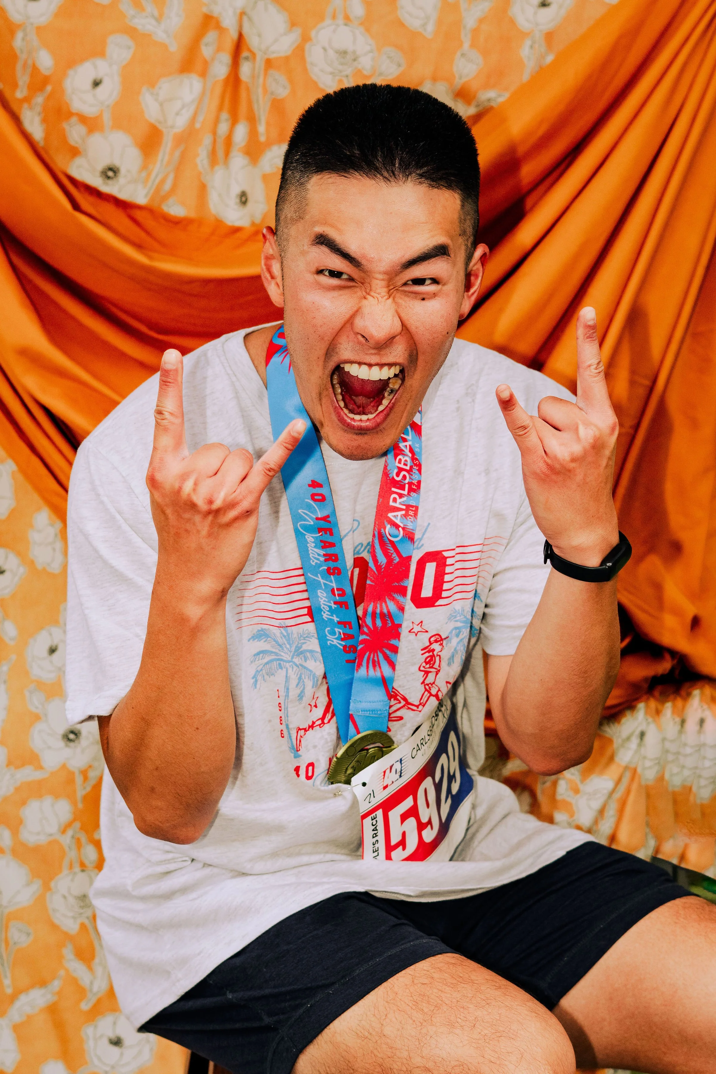 A young man with short black hair is sitting on a floral bench, smiling widely with his mouth open, making rock and roll hand gestures with both hands. He is wearing a white T-shirt with red and blue design, a race bib, a blue and red medal around hi