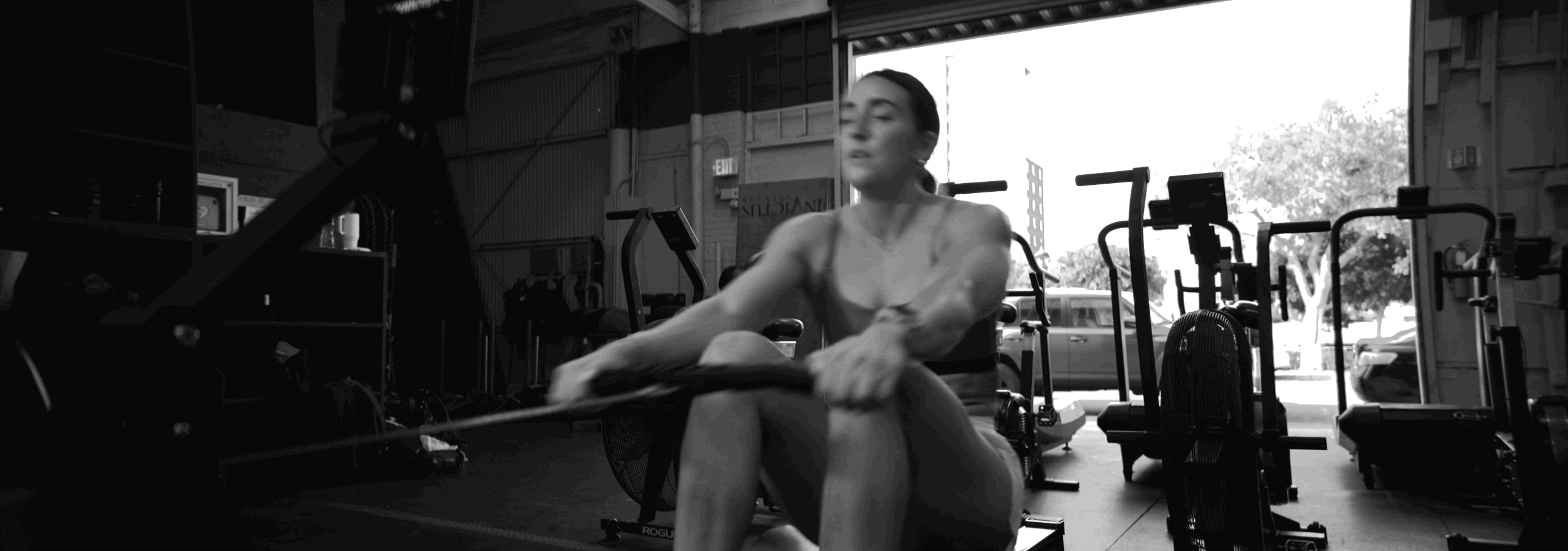 A woman working out with a rowing machine inside a gym, with other workout equipment visible in the background and cars outside the open garage door.