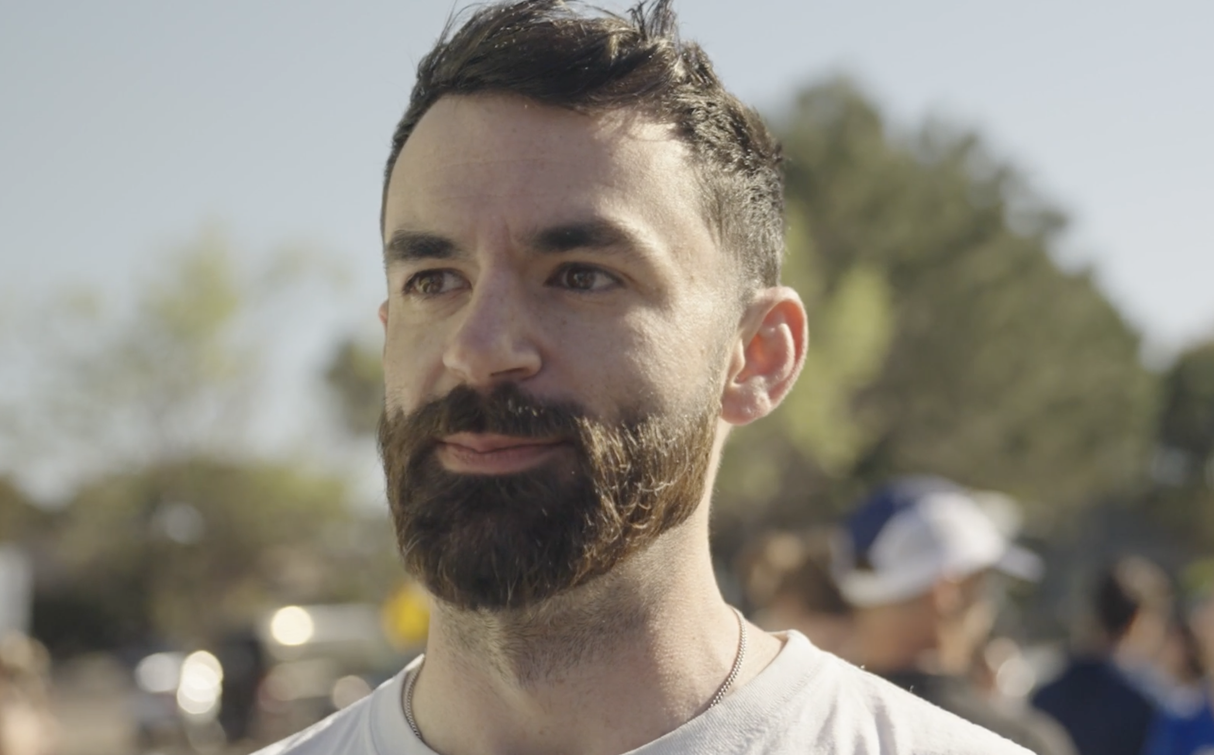 Close-up of a young man with dark hair and a beard, outdoors on a sunny day with blurred trees in the background.