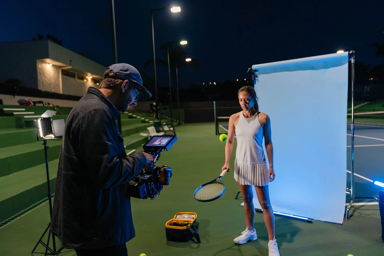 A female tennis player is holding a tennis racket and a tennis ball, standing in front of a light blue backdrop during a nighttime photoshoot on a tennis court with artificial lighting.