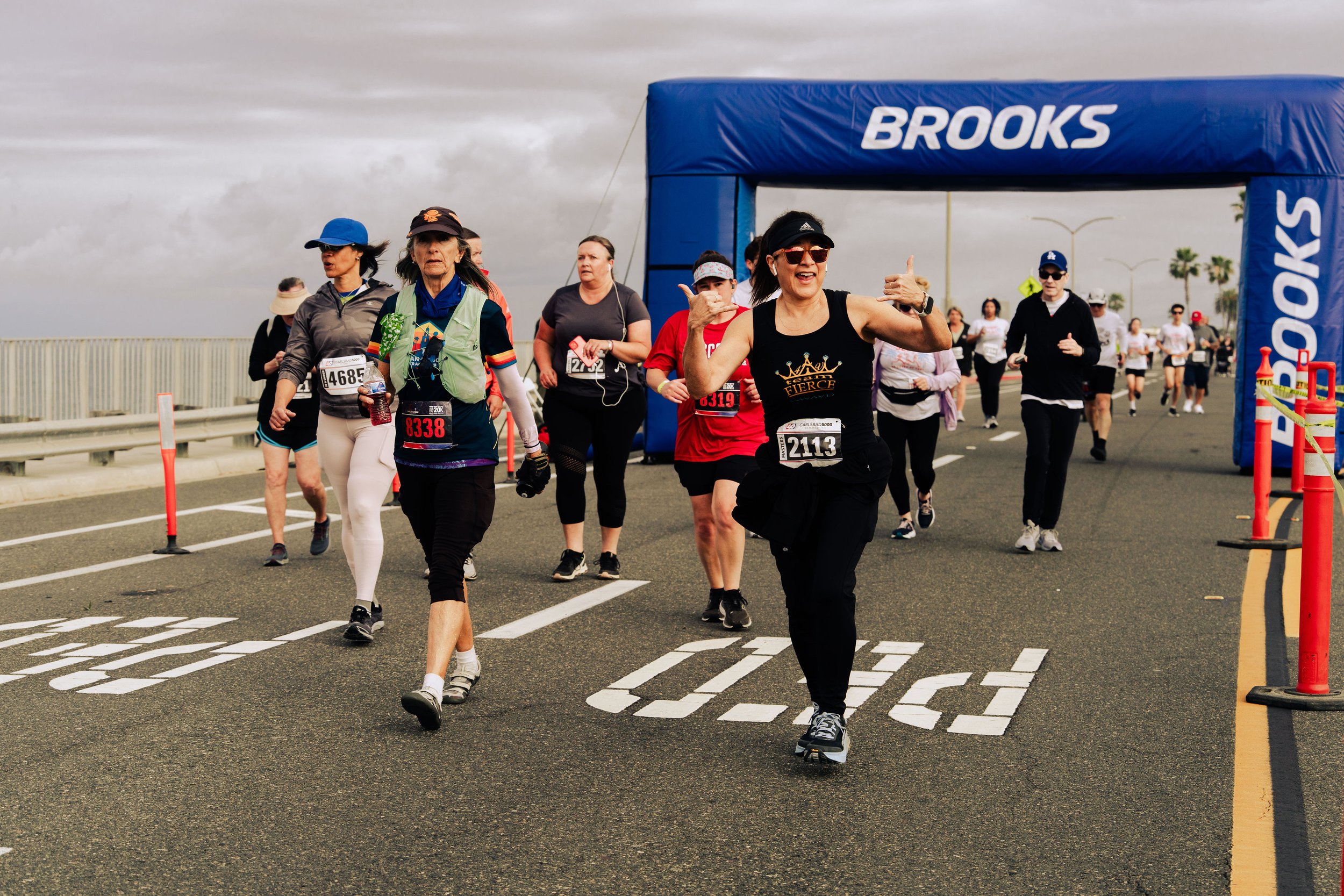 People participating in a marathon or running event, crossing the starting line under a blue archway with the word 'Brooks' on it, on an overcast day.