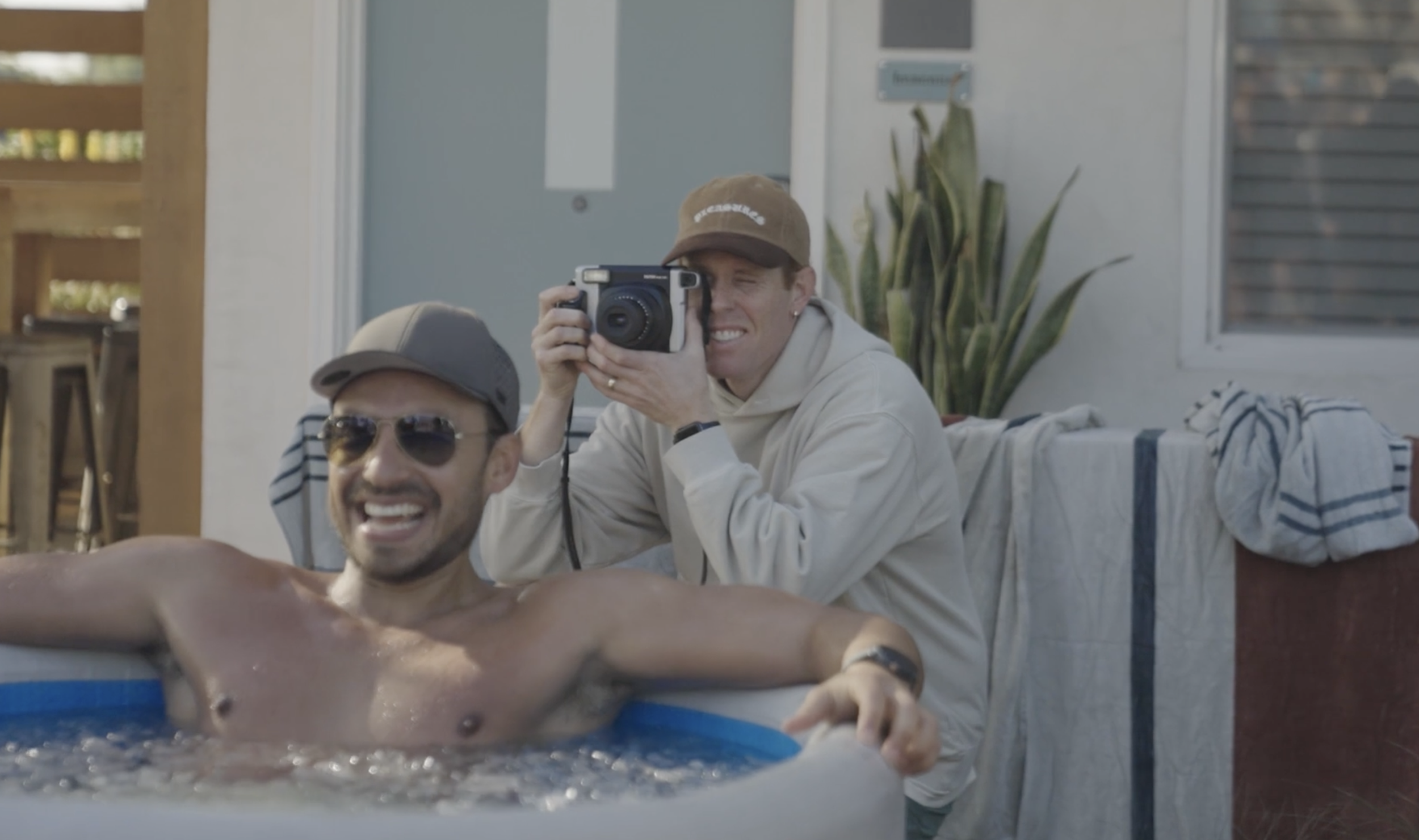 Two men are enjoying a day by the pool, with one of them taking a photo of the other who is lying in the pool.