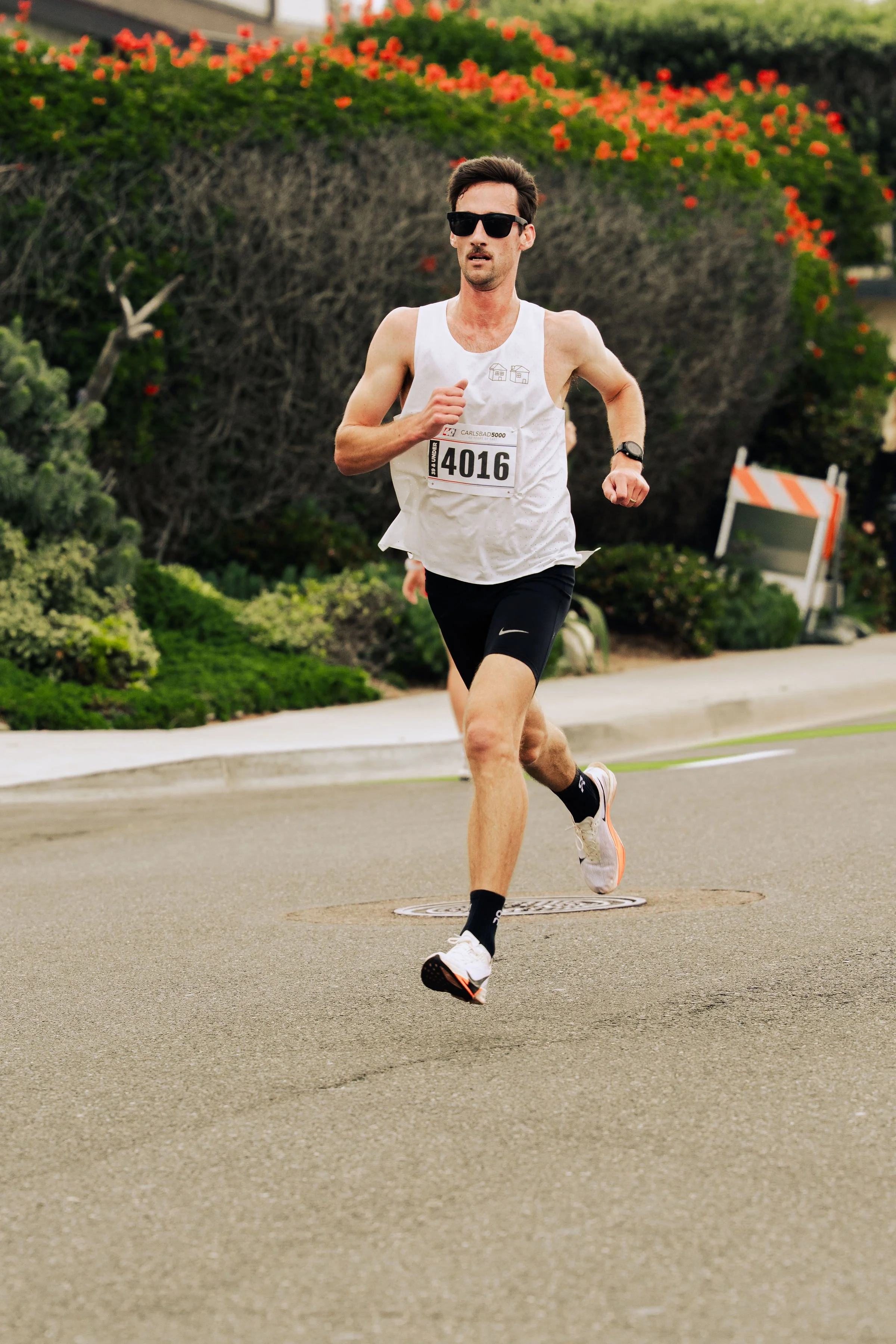 A man wearing sunglasses, a white tank top with a race bib number 4016, black shorts, and running shoes is running on a street during a race event, with greenery and flowers in the background.