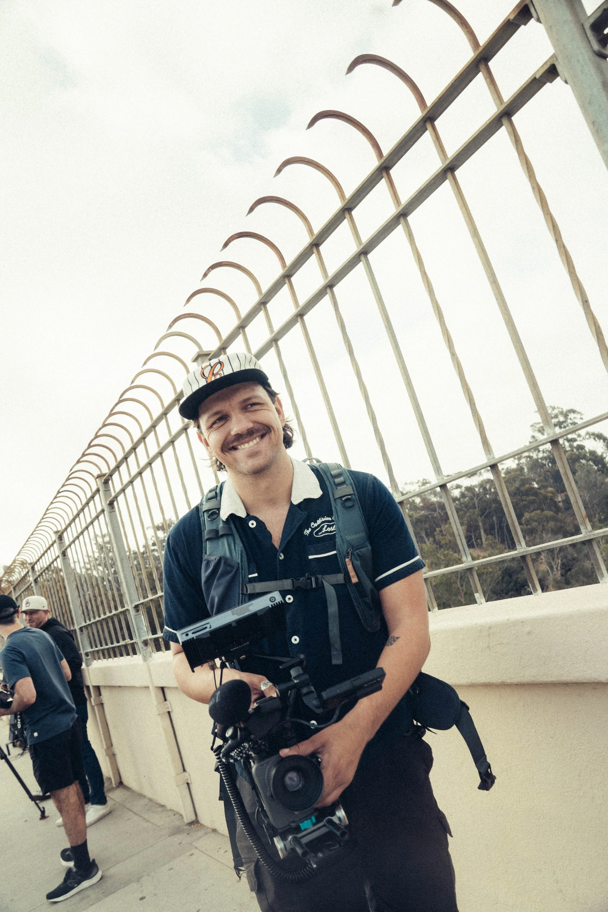 A young man smiling and holding a camera in front of a metal fence, with several other people in the background.