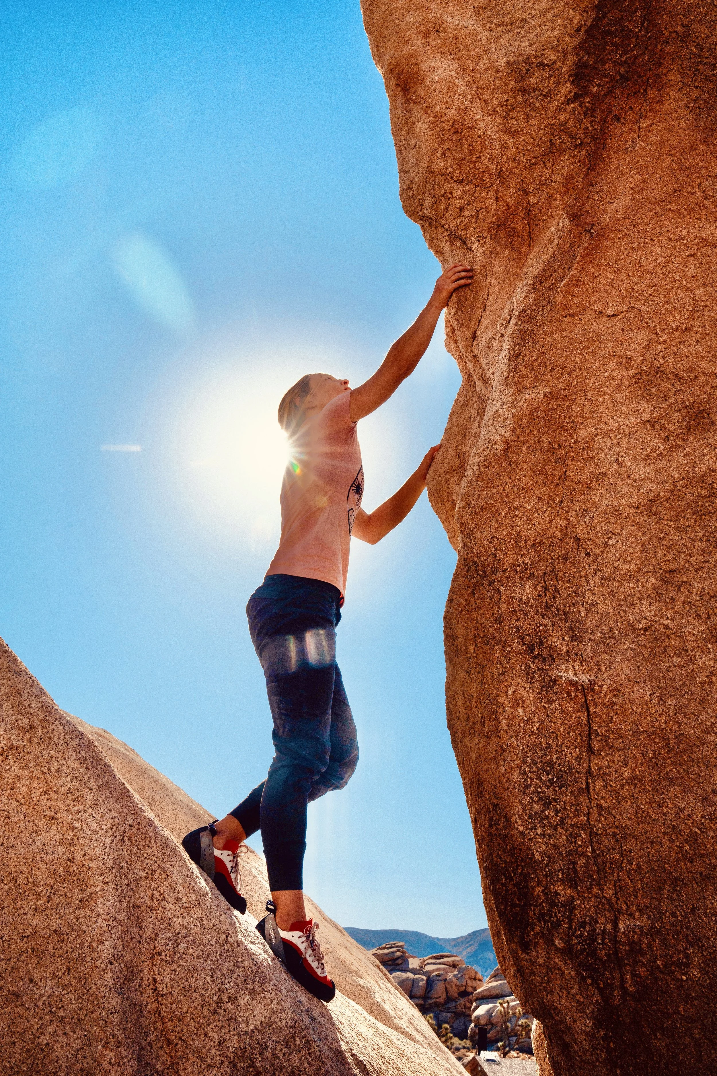 A woman rock climbing on a large reddish-brown rock formation under a bright blue sky with the sun shining behind her.