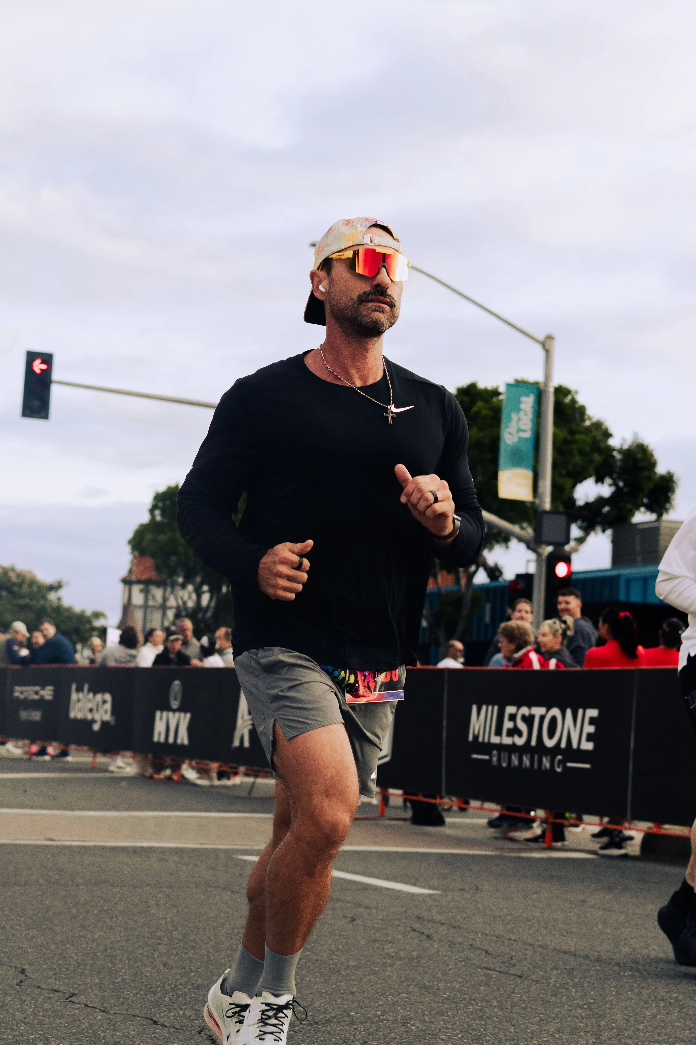A man running in a street race, wearing sunglasses, a black shirt, gray shorts, white sneakers, a beige cap backward, a cross necklace, and wireless earbuds, with a crowd and race banners in the background.