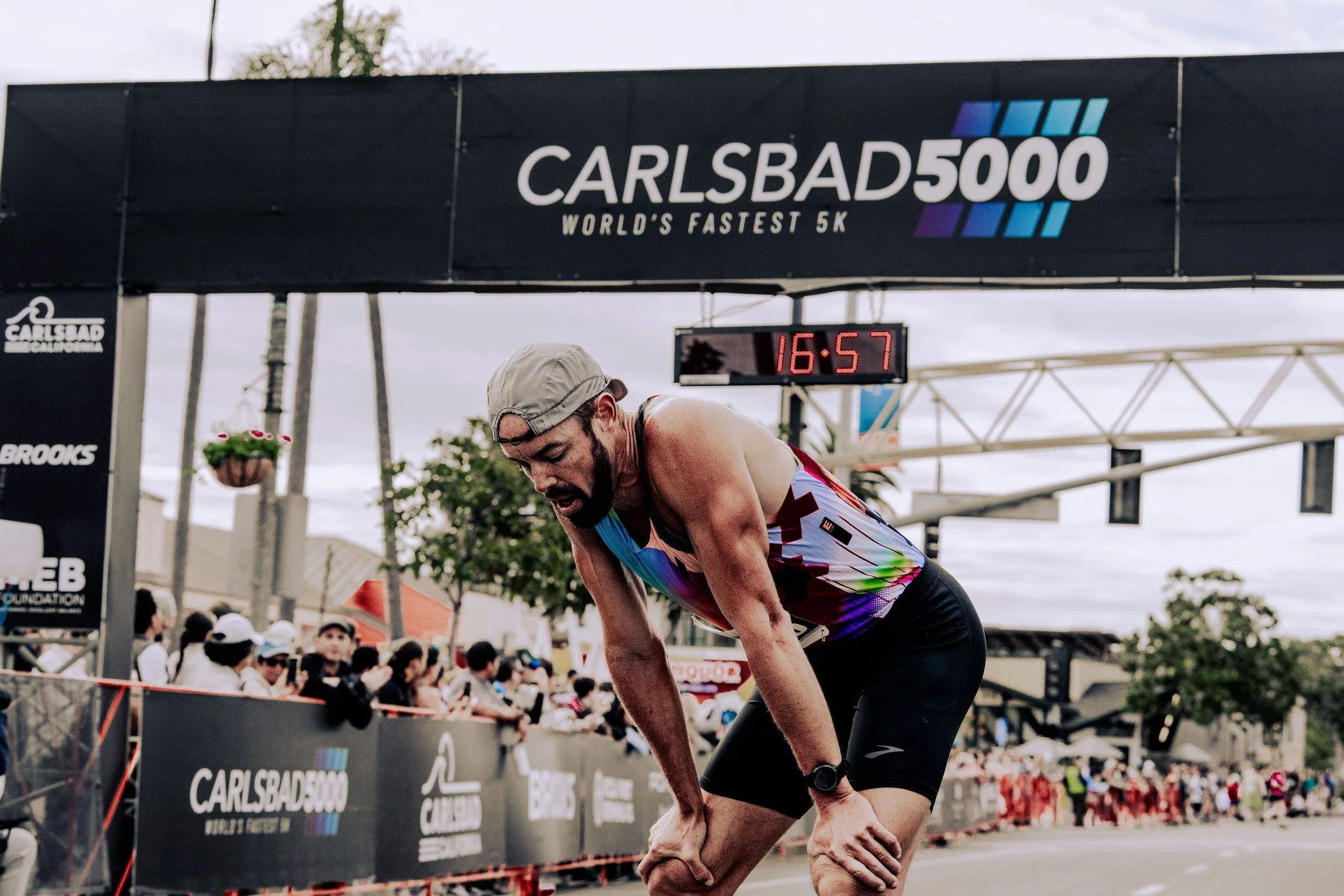 A male runner in colorful athletic gear crosses the finish line at the Carlsbad 5000, World's Fastest 5K, with a digital clock showing 16 minutes and 57 seconds in the background, surrounded by spectators.