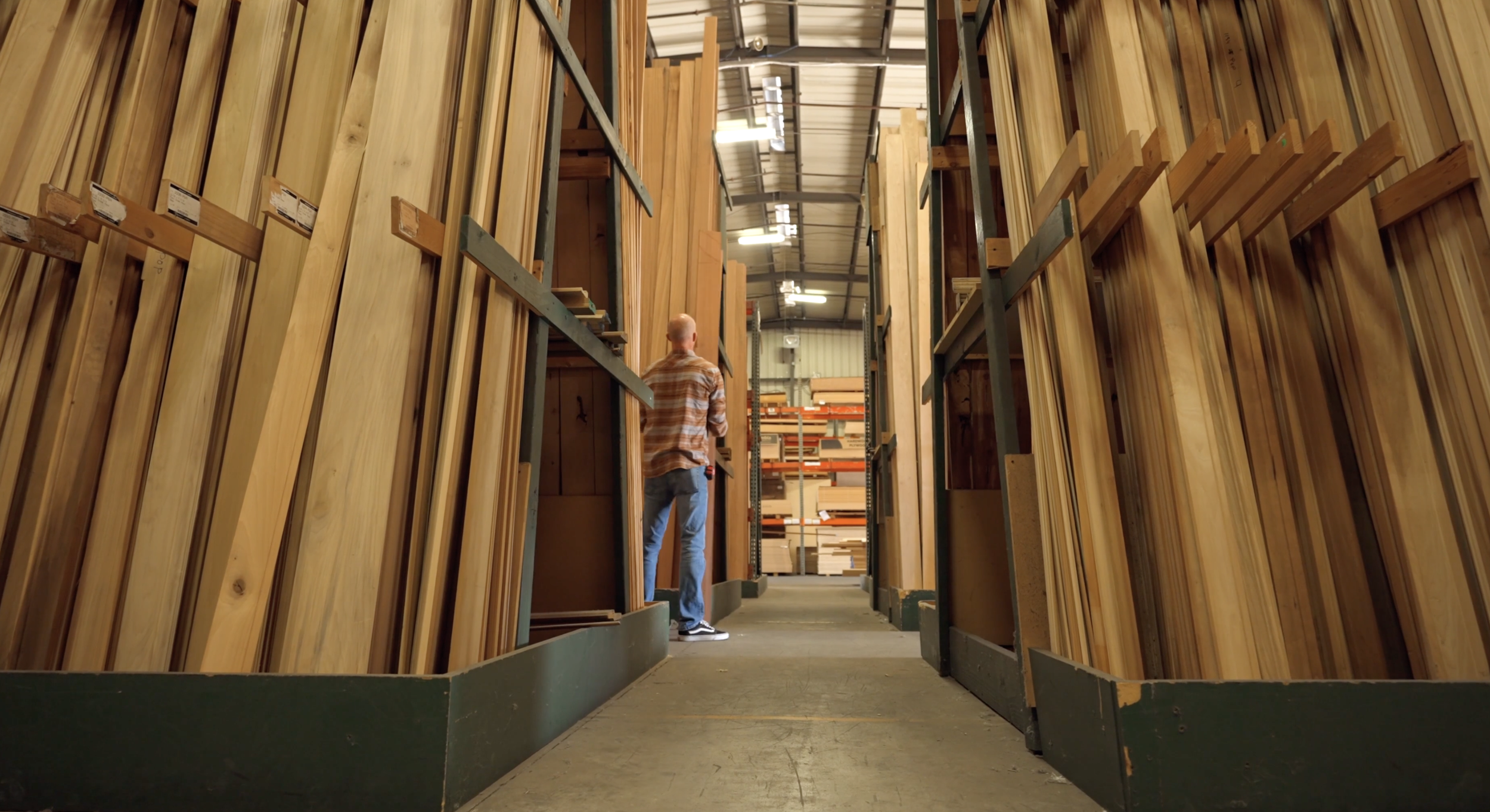 Person shopping for wood planks in a hardware store aisle with high shelves filled with various types of lumber.