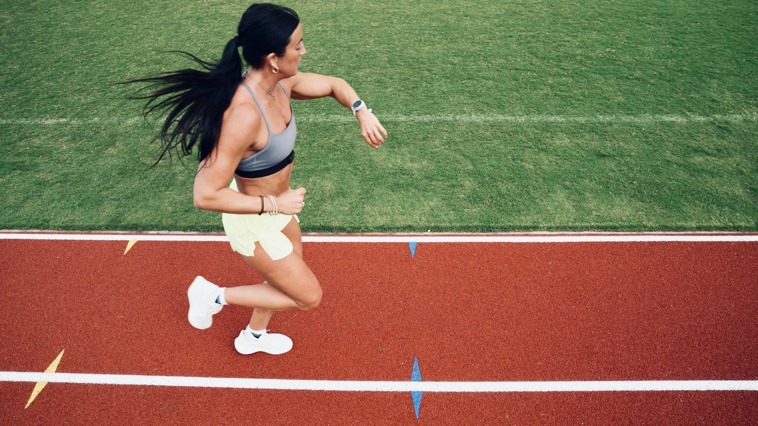 A woman running on an outdoor track, wearing sportswear, with grass field in the background.