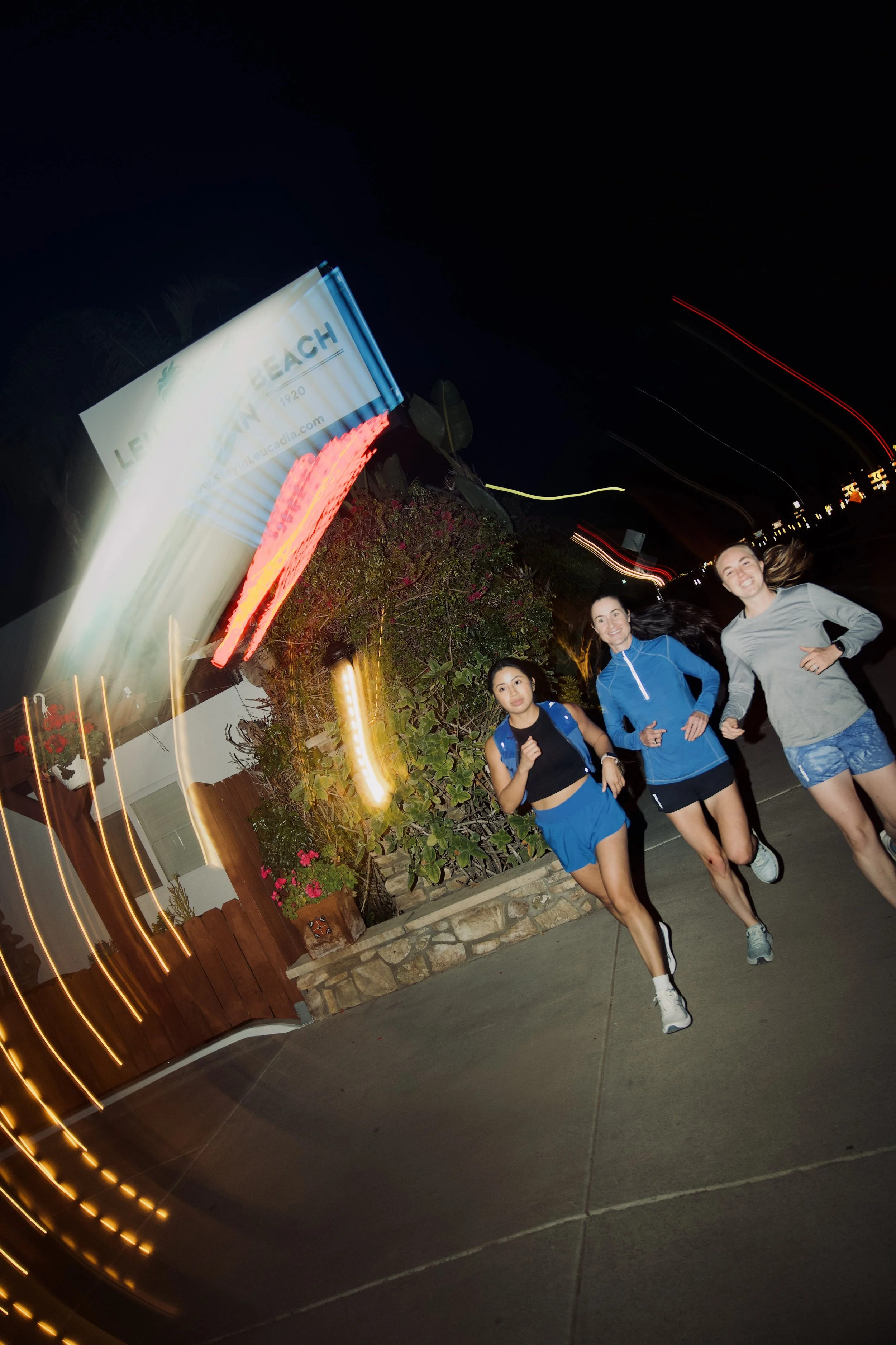 Three women jogging on a sidewalk at night near a lit sign and plants, with light streaks in the sky from camera movement.