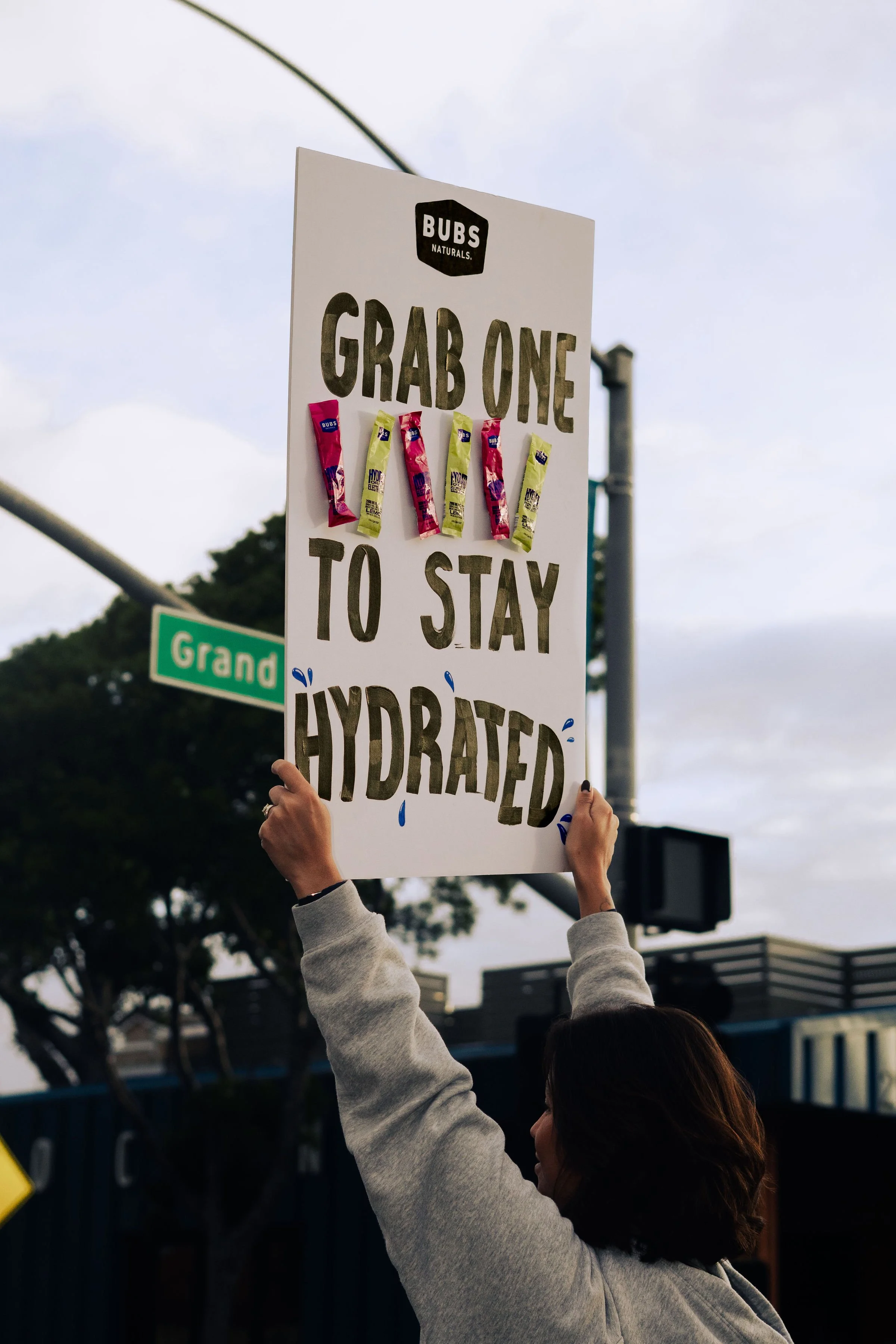 Person holding a protest sign that reads 'Grab one to stay hydrated' with small hydration packet stickers, outdoors near a street sign.