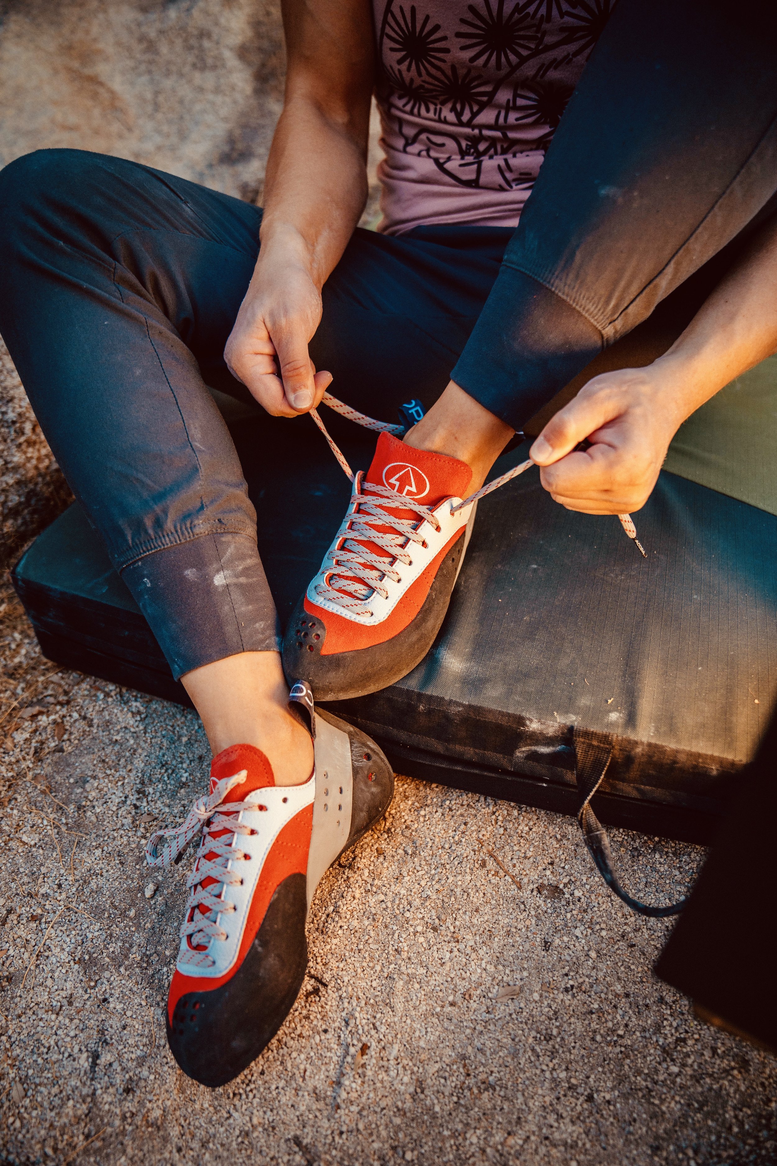 A person sitting on a portable climbing crash pad on a sandy ground, tying the laces of their climbing shoes. The person is wearing black pants, a pink shirt with a black pattern, and a black wristband. The climbing shoes are white with orange and bl