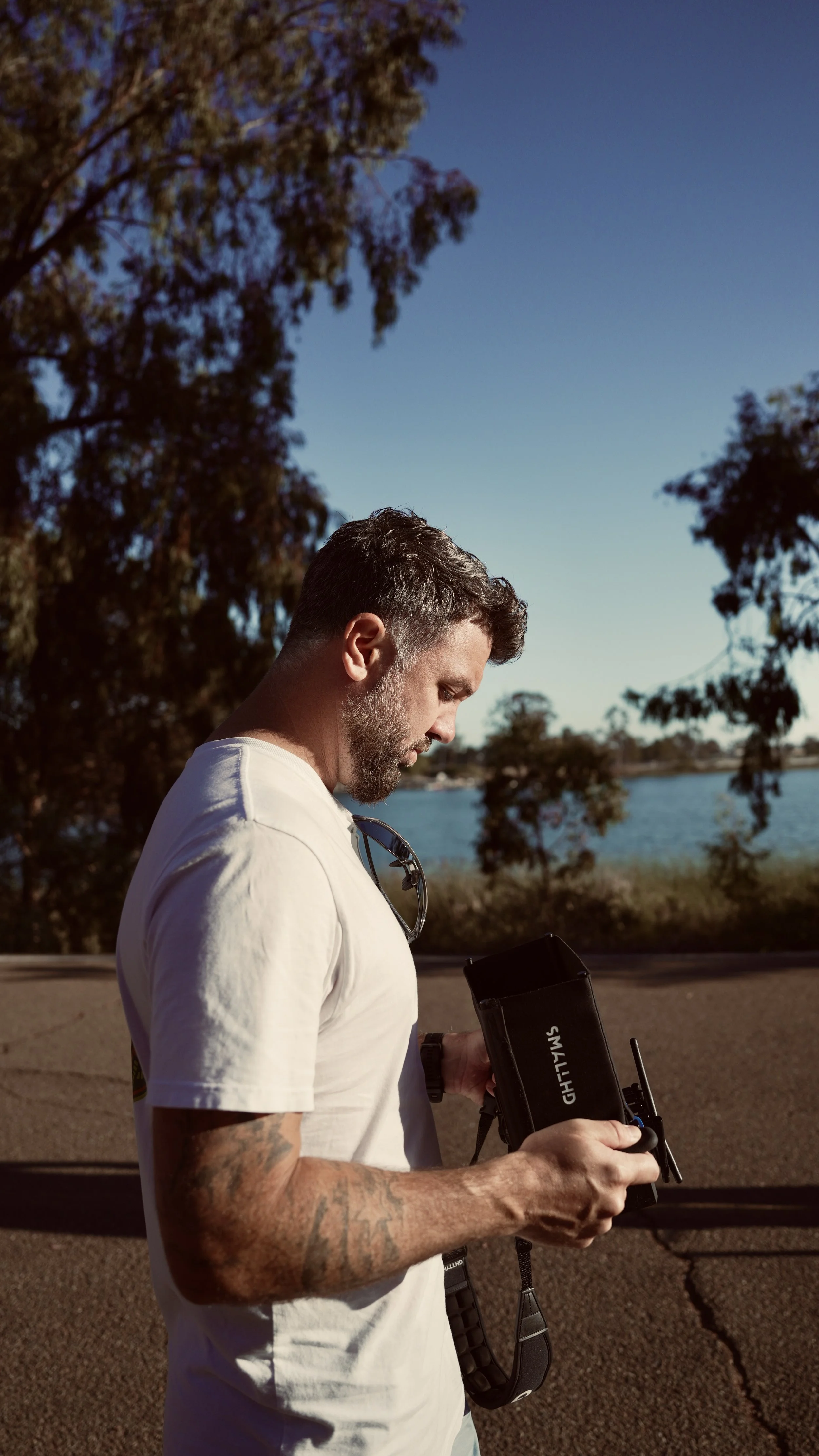 A man with short dark hair and tattoos on his arms, wearing a white t-shirt, is holding and operating a small handheld camera or monitor outdoors near a body of water. There are trees and clear blue sky in the background.