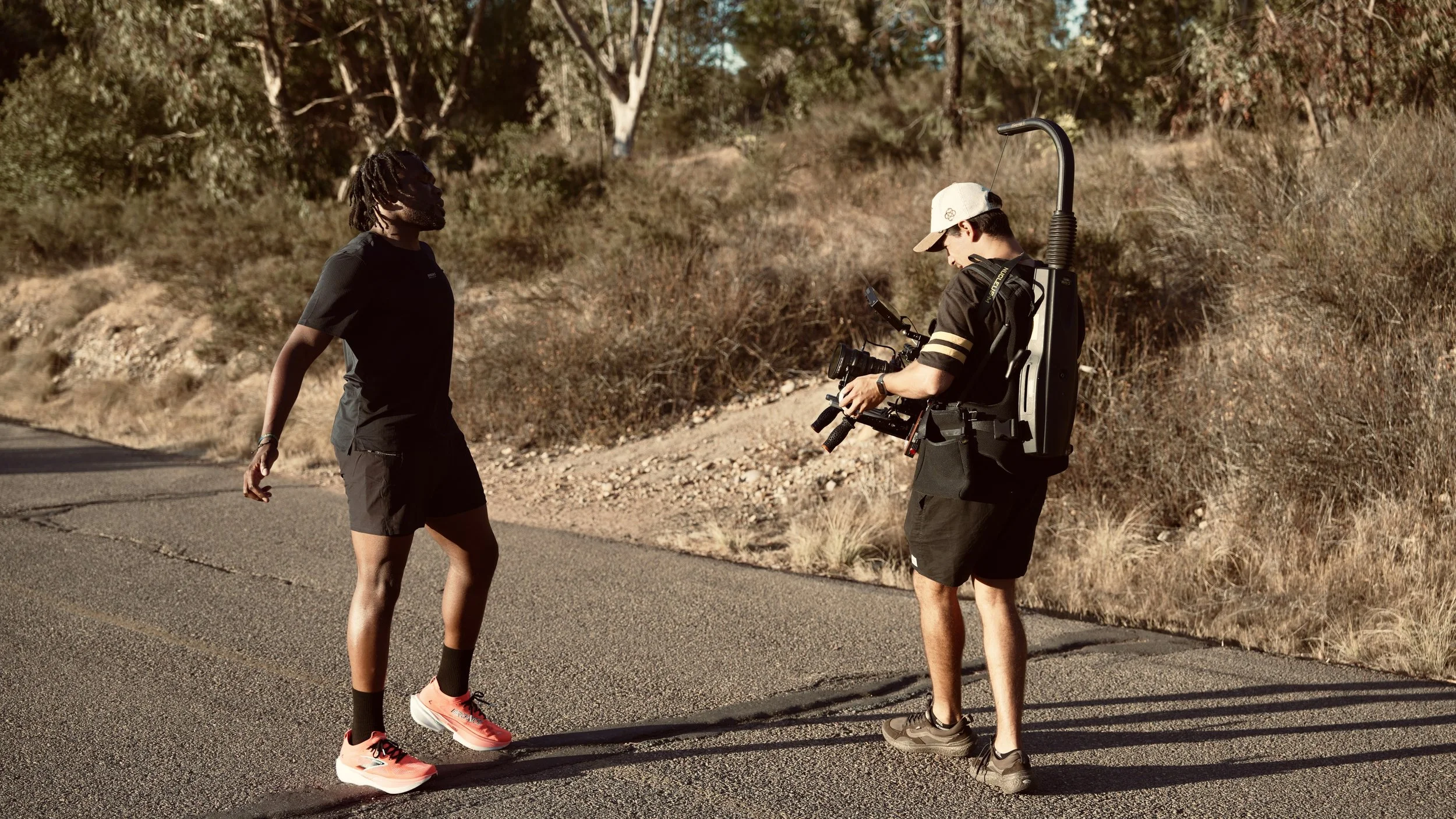 Two people having a conversation on the side of a rural road, one with a camera backpack and the other dressed in athletic gear, with trees and dry brush in the background.