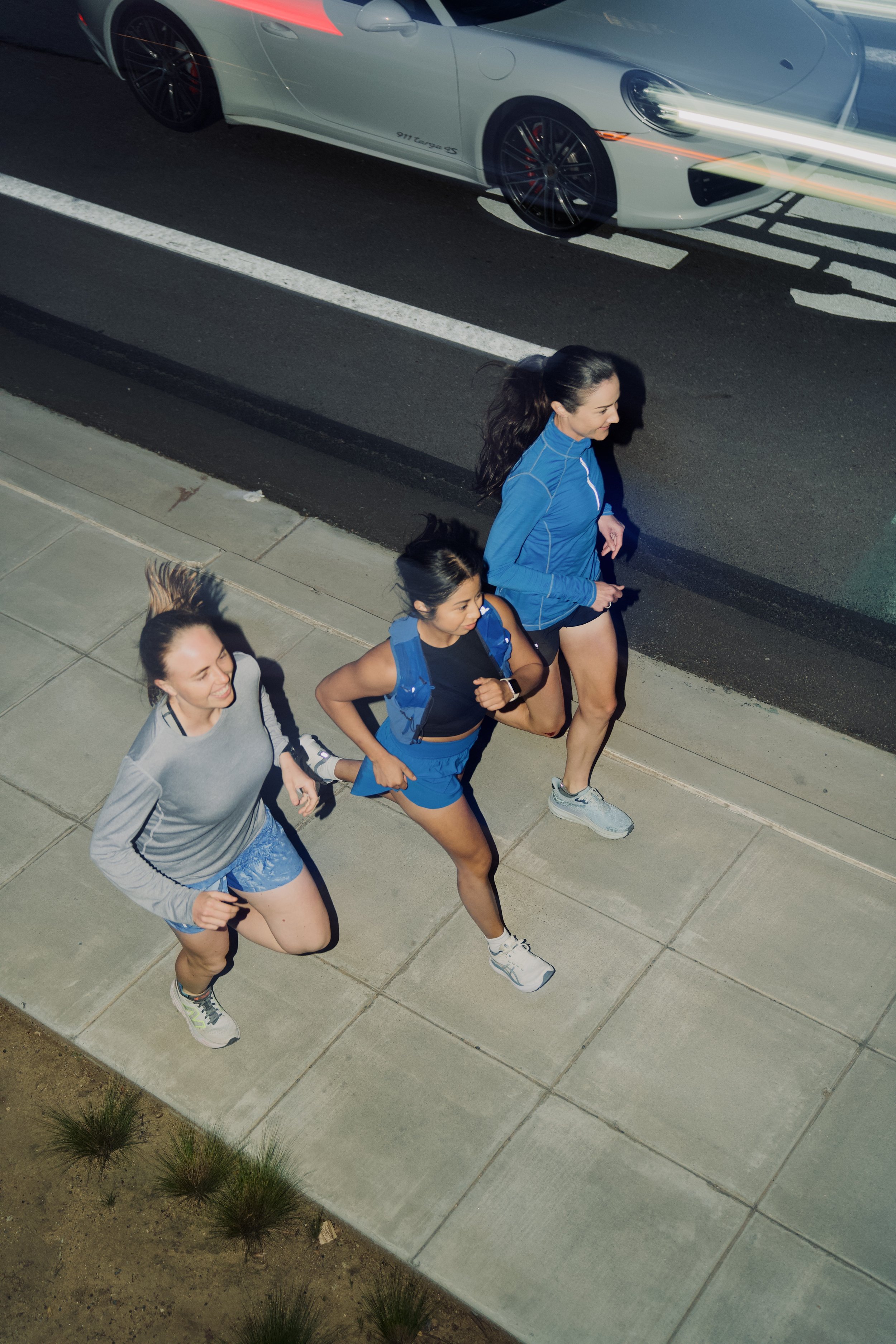 Three women in running gear jogging on a sidewalk, viewed from above, with a white sports car parked on the street nearby.