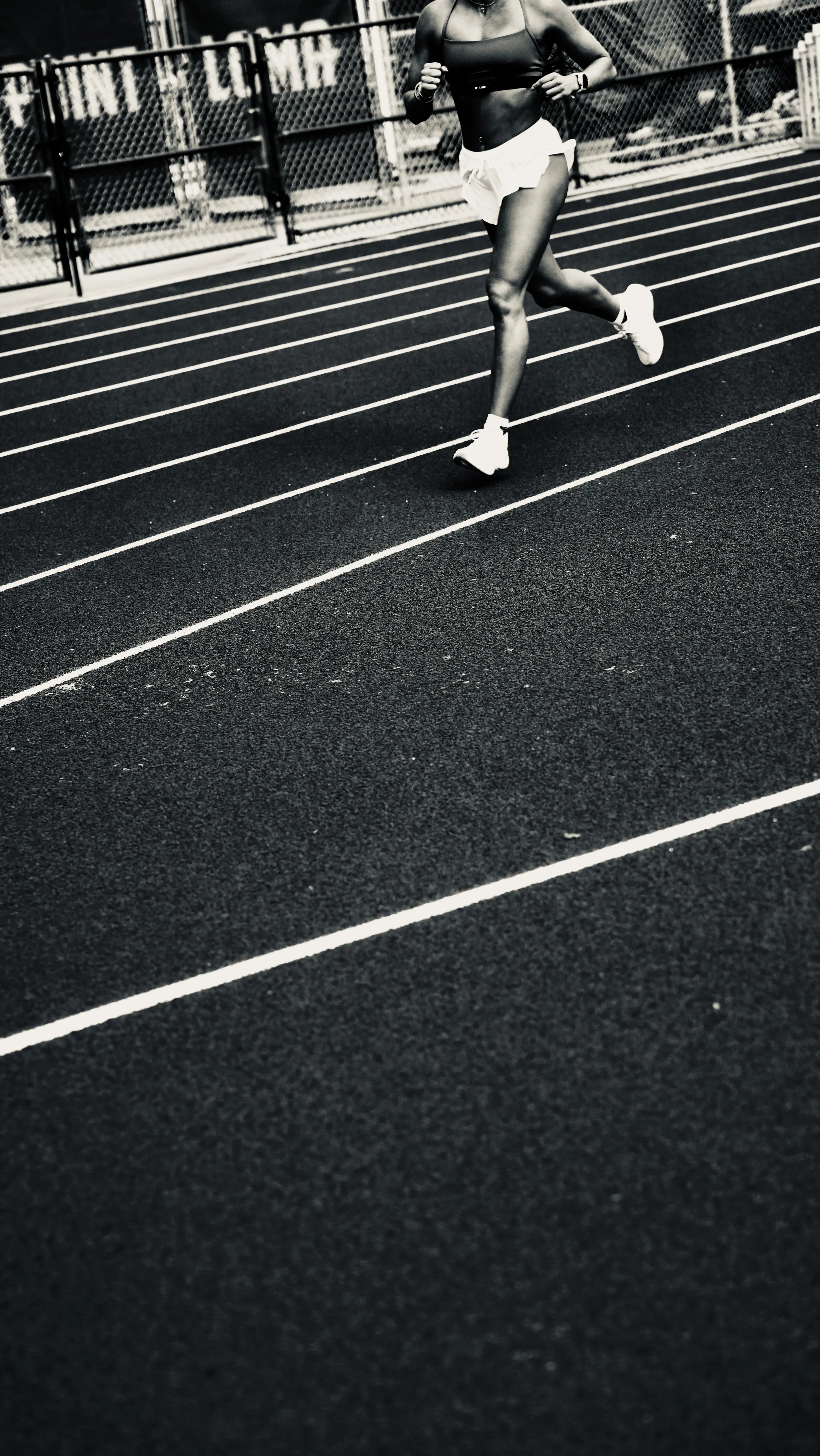 A woman running on a black outdoor track with white lane lines, wearing a sports bra, shorts, and running shoes.