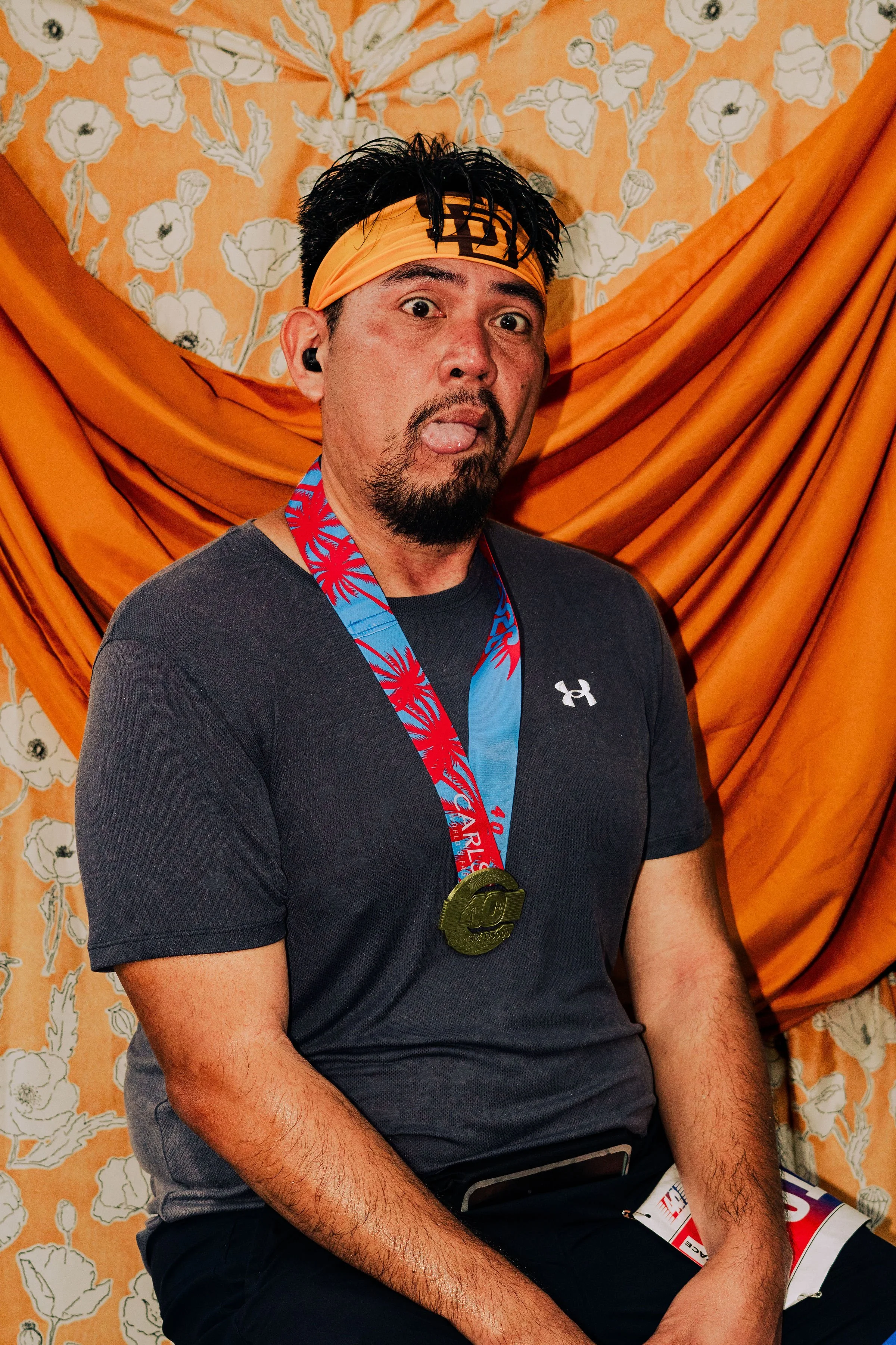 A man with black hair, a goatee, and wearing a yellow headband sticks out his tongue. He has a medal around his neck and is sitting in front of orange drapes and floral wallpaper.