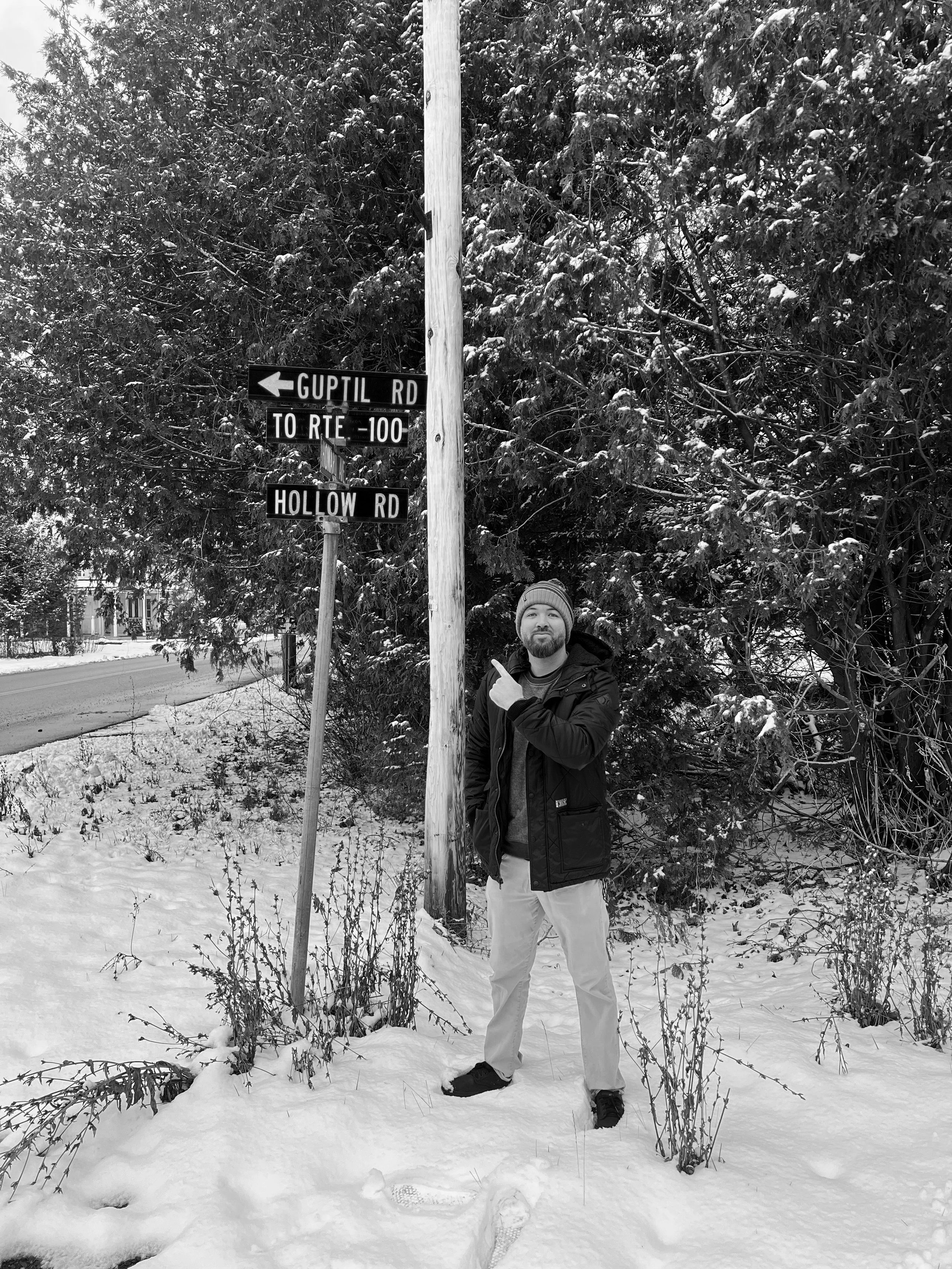 A man standing in a snowy landscape pointing at a street sign that reads 'GUPTIL RD' and 'HOLLOW RD' with a background of snow-covered trees.