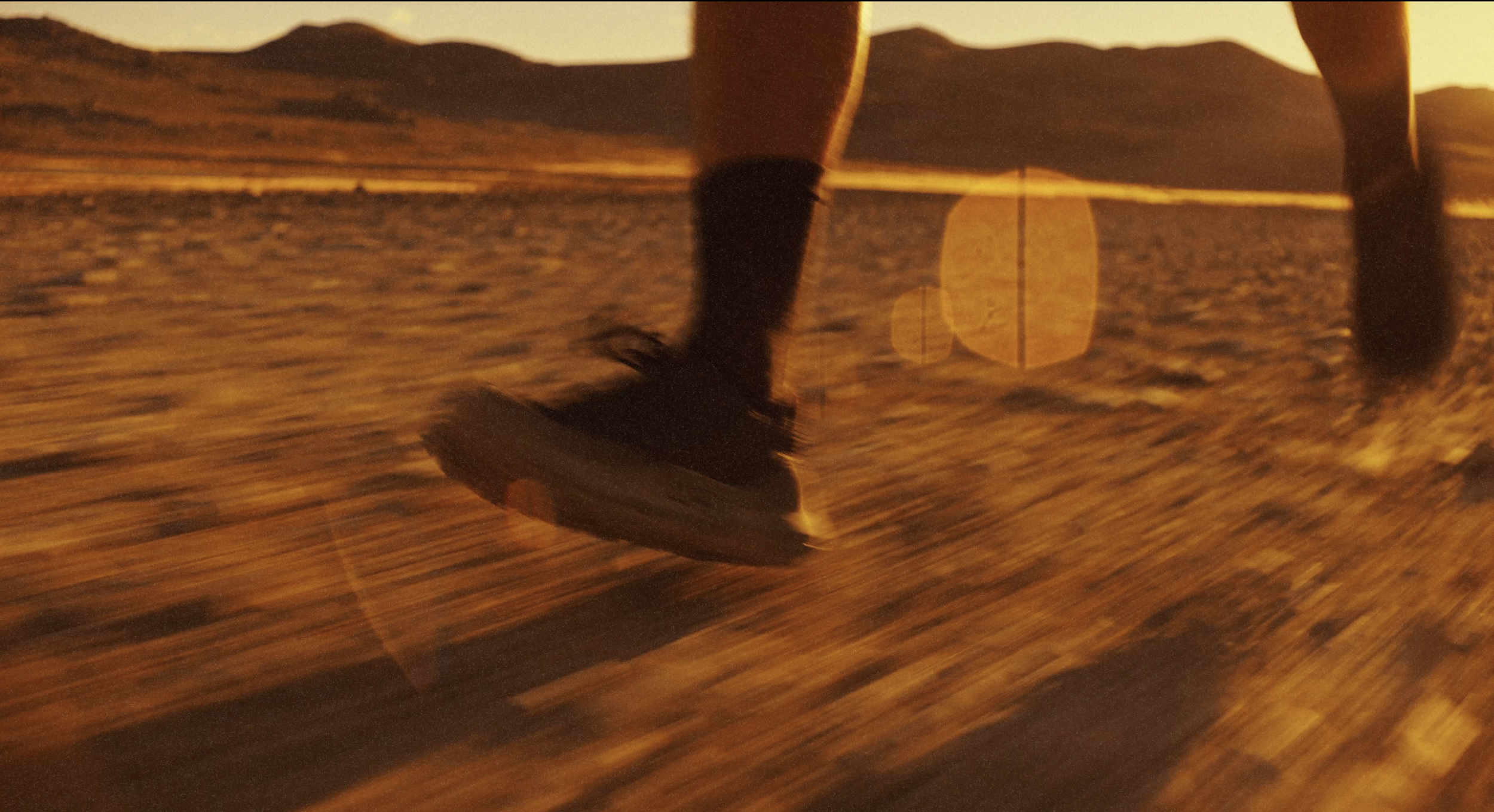 Close-up of a skateboard wheel and deck, with a person's legs visible, on a dirt surface during sunset with distant mountains in the background.
