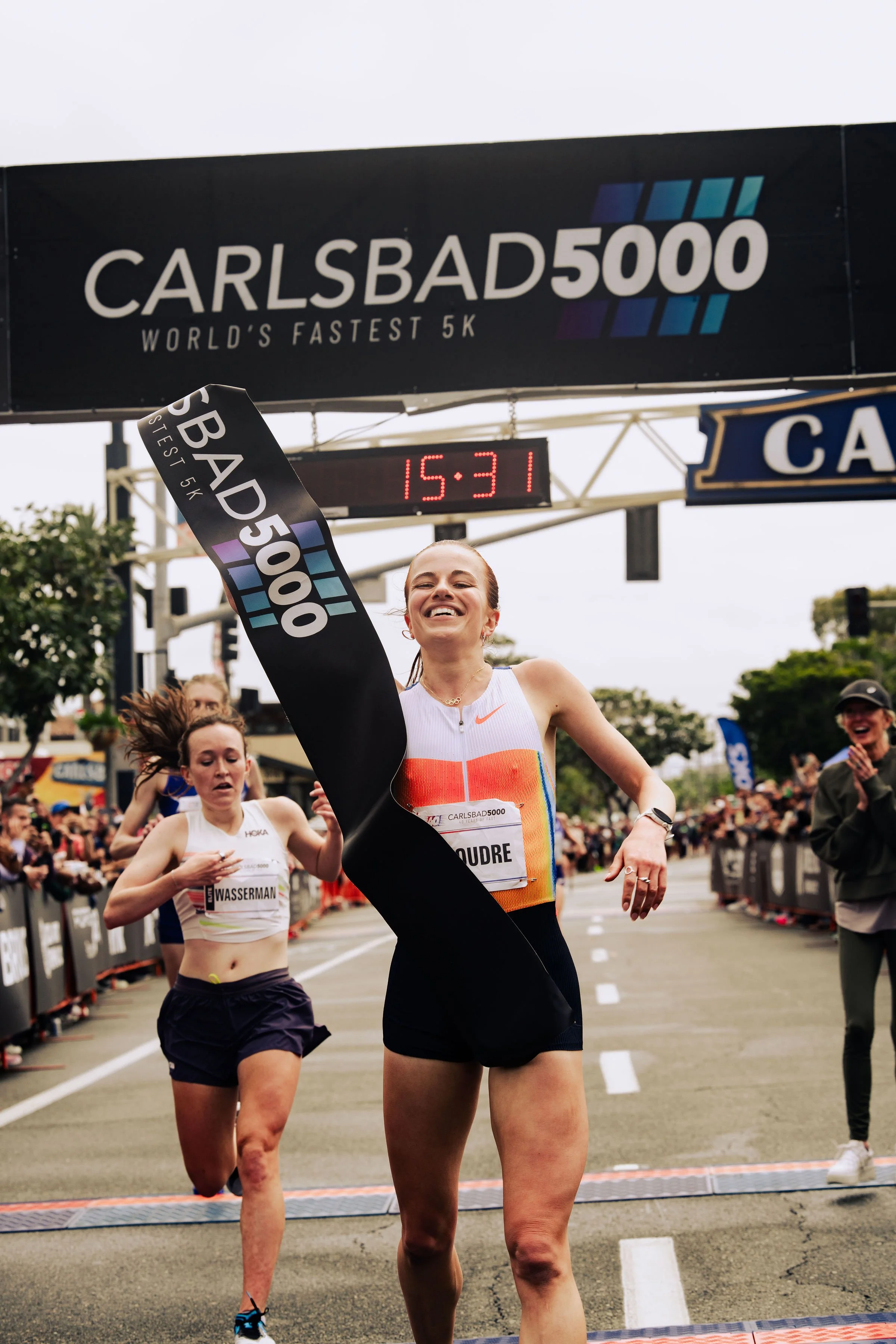 A female runner crossing the finish line at the Carlsbad 5000 race, smiling with her arm raised in celebration. Other runners and spectators are visible in the background.