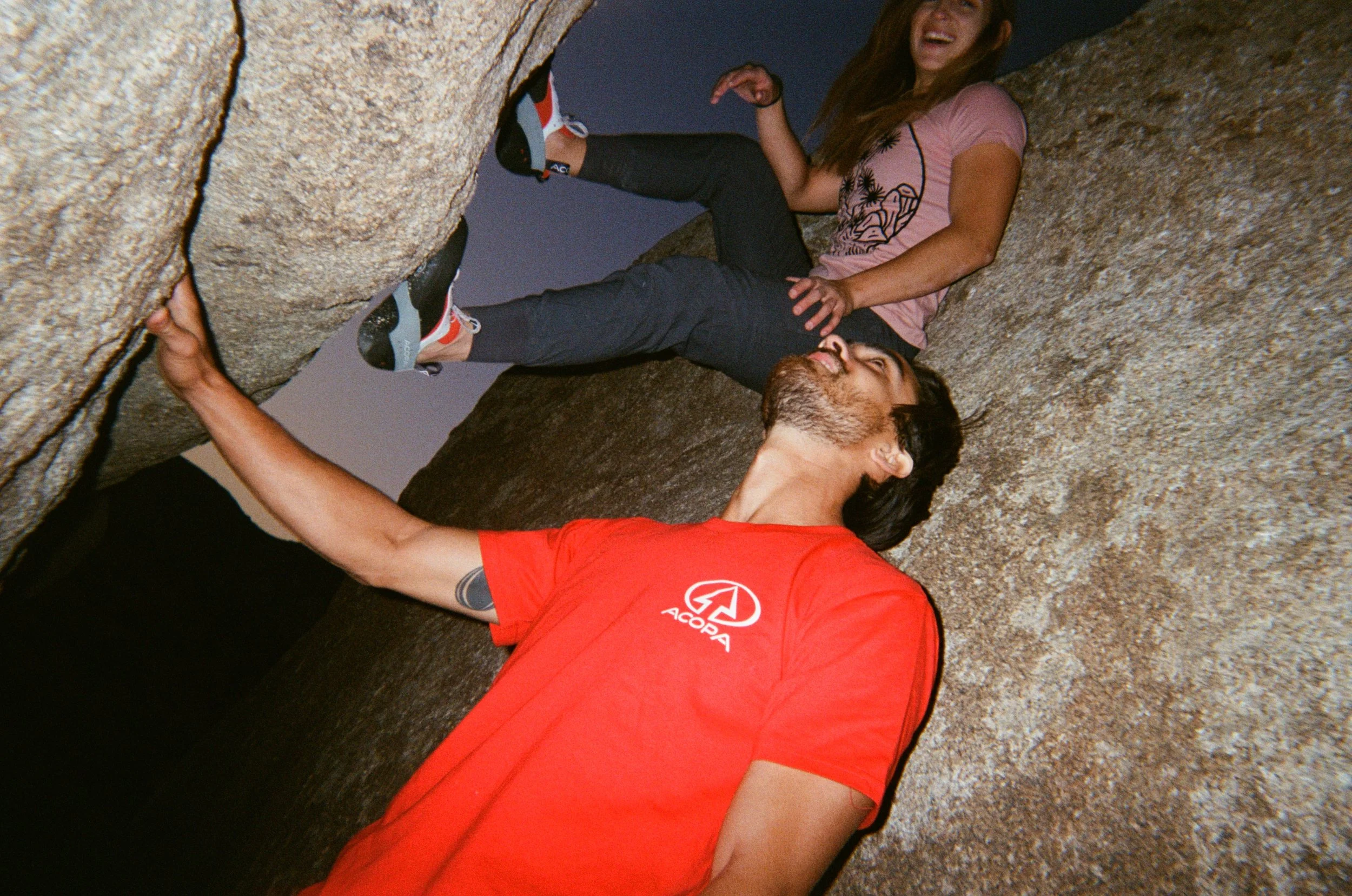 A man and woman appear to be rock climbing on a large granite rock formation, with the woman sitting and the man lying on the ground, both smiling and looking at each other.
