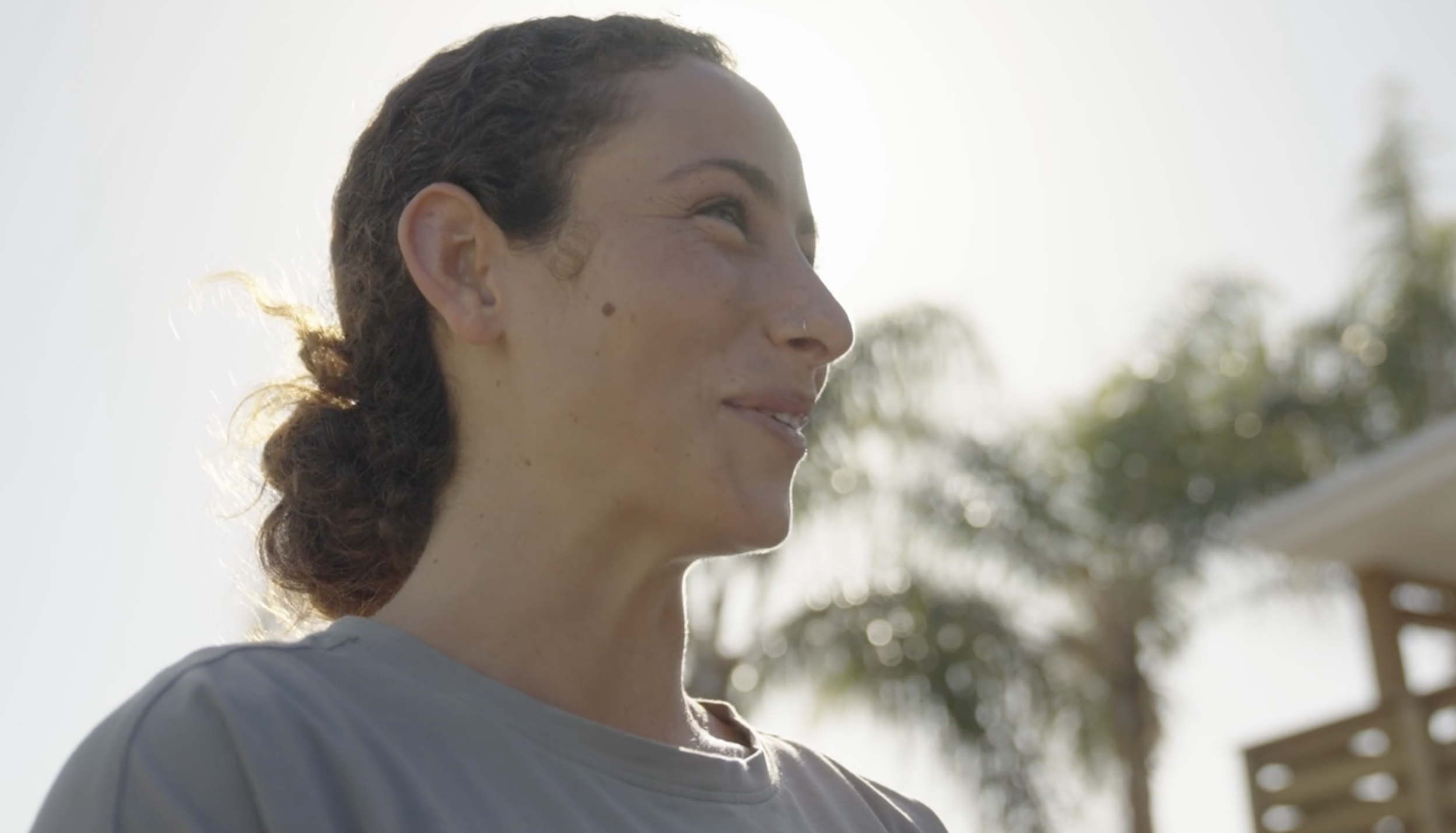 A woman with curly hair and freckles smiling outdoors with sunlight behind her and trees in the background.
