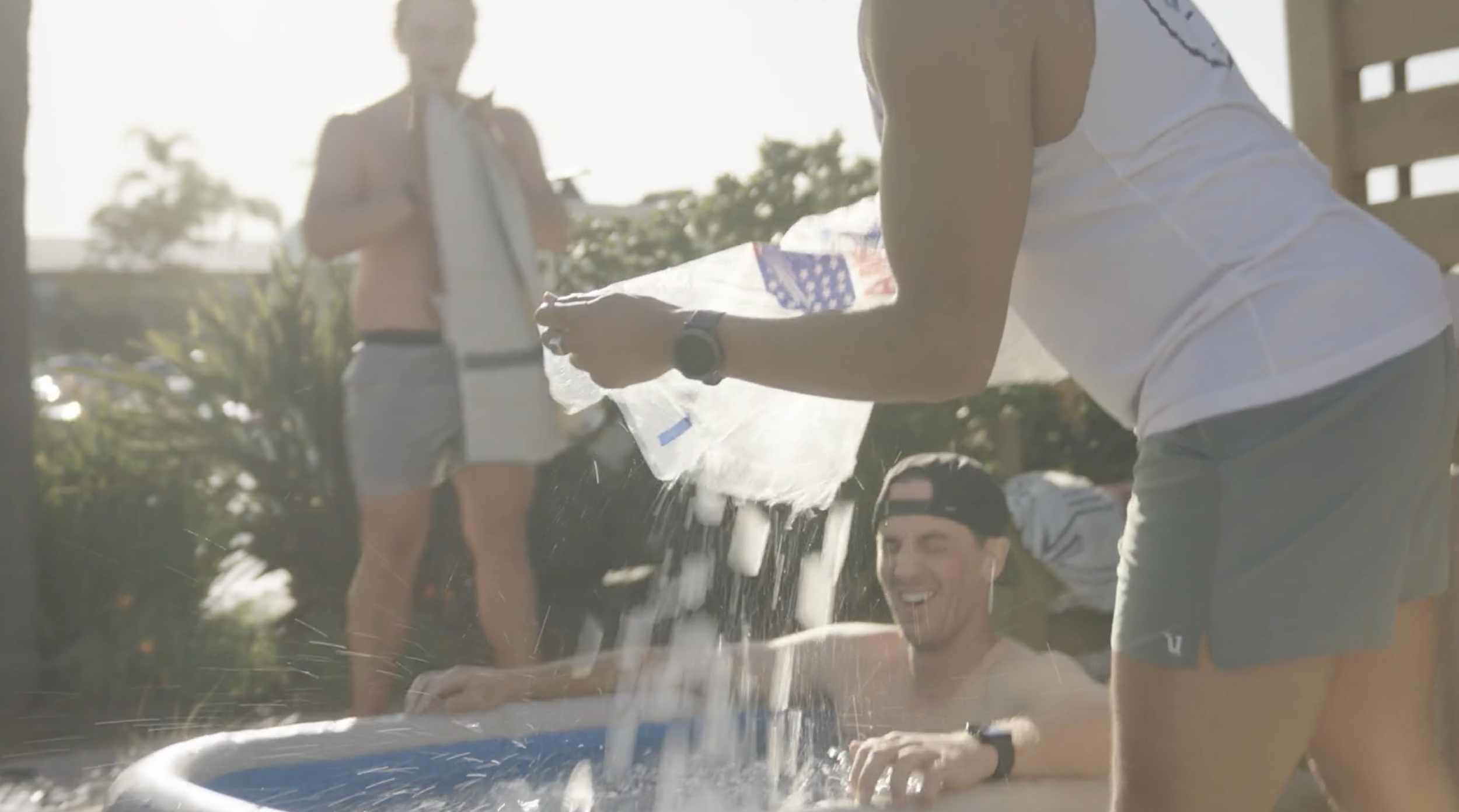 Two people smiling and playing with water outside on a sunny day, with a man kneeling and holding a plastic bag, and a woman standing in the background.