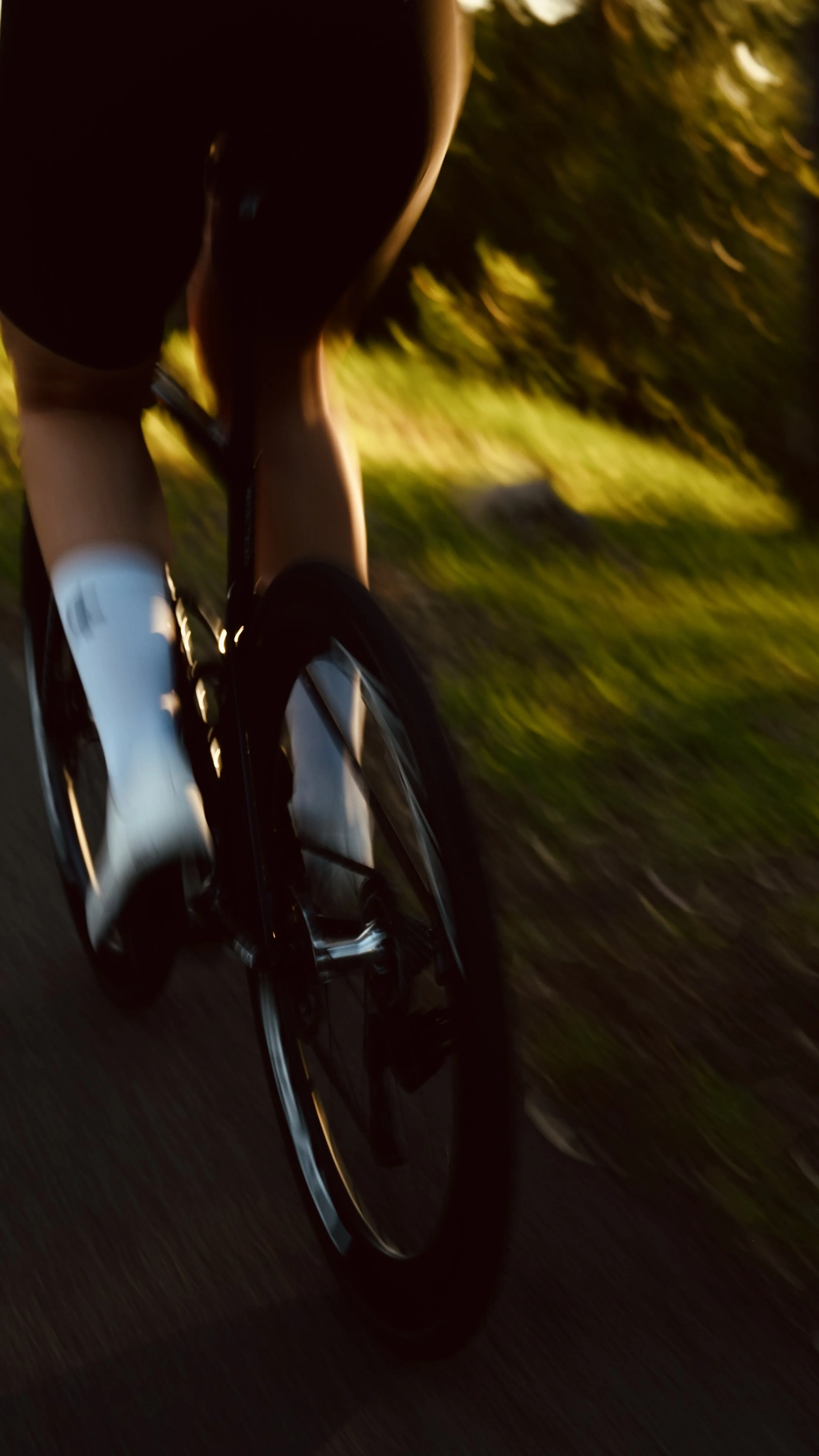 Close-up of a person riding a bicycle on a road at sunset, with the shot taken from behind and slightly below the rider, capturing the motion blur of the background and the bicycle.