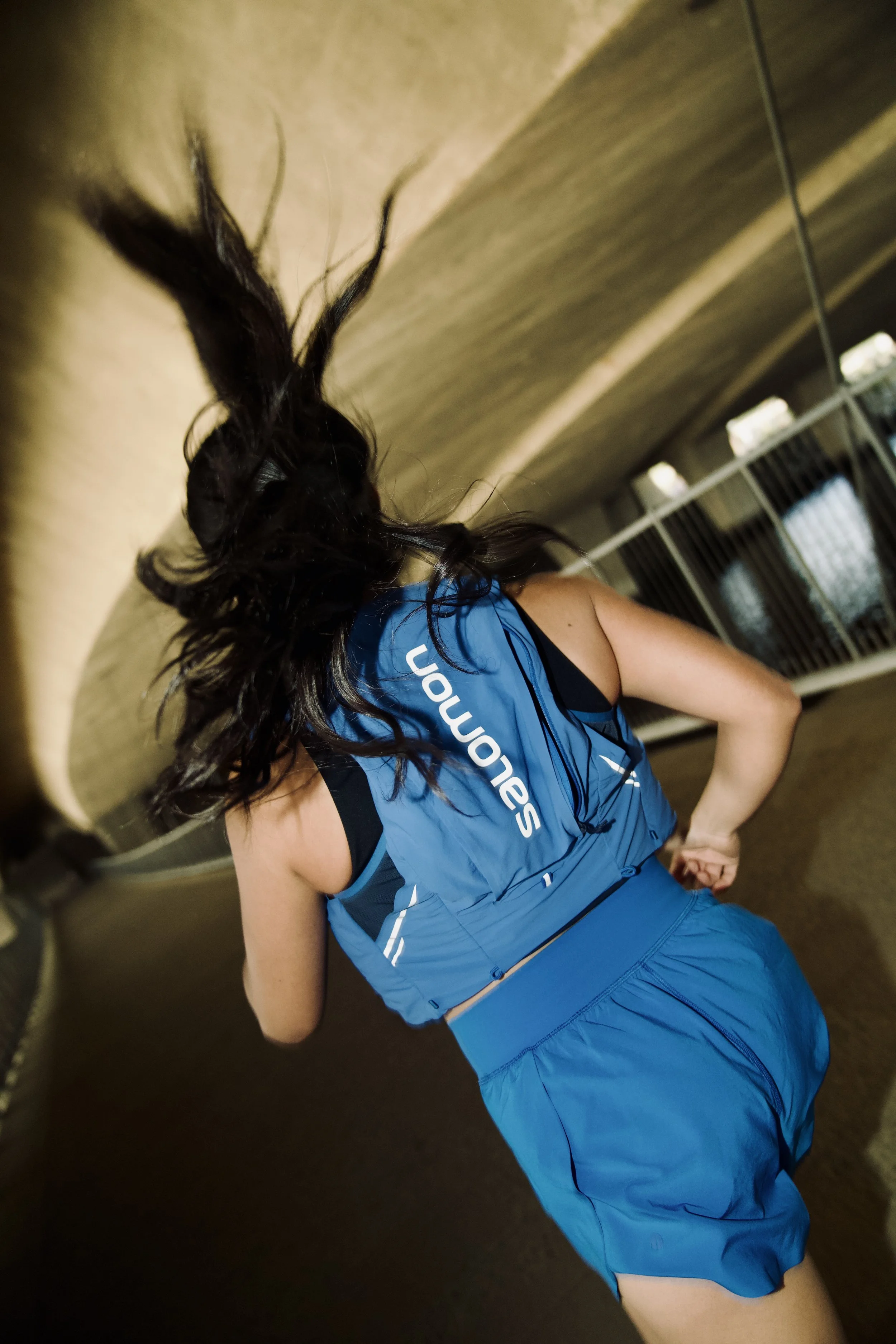 A person wearing a blue sports uniform with the word 'SENATORS' on it, standing with their back to the camera in an indoor sports facility, with long dark hair, and with a railing and windows in the background.