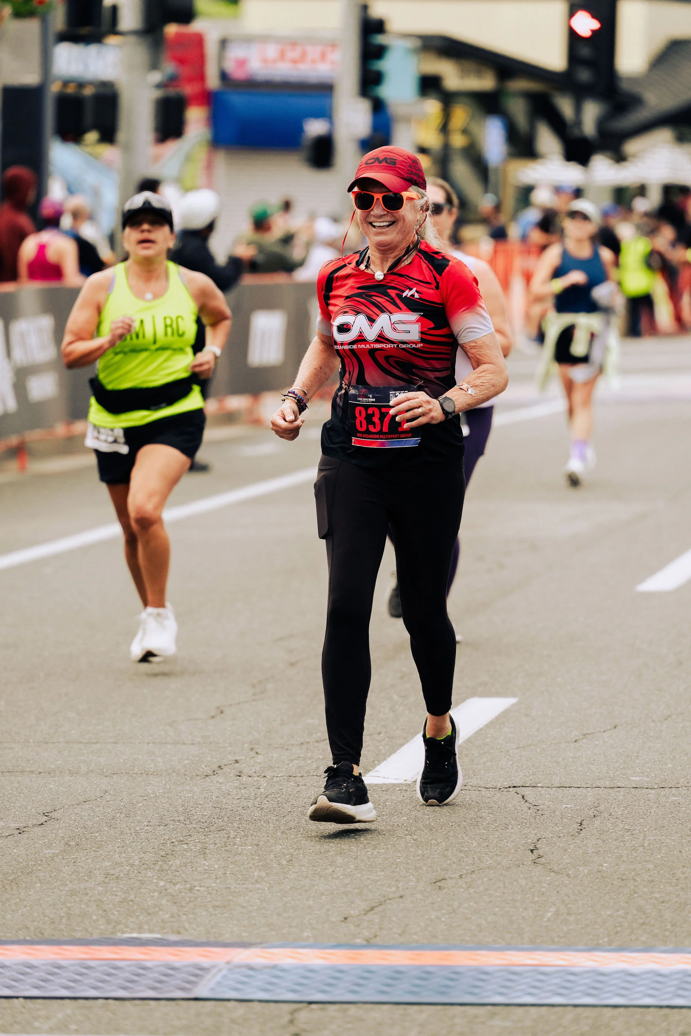 A woman smiling while running in a marathon, wearing a red cap, sunglasses, and a red and black athletic shirt with the logo 'OMG'. She has a race bib with the number 837. In the background, other runners and spectators can be seen on the street.