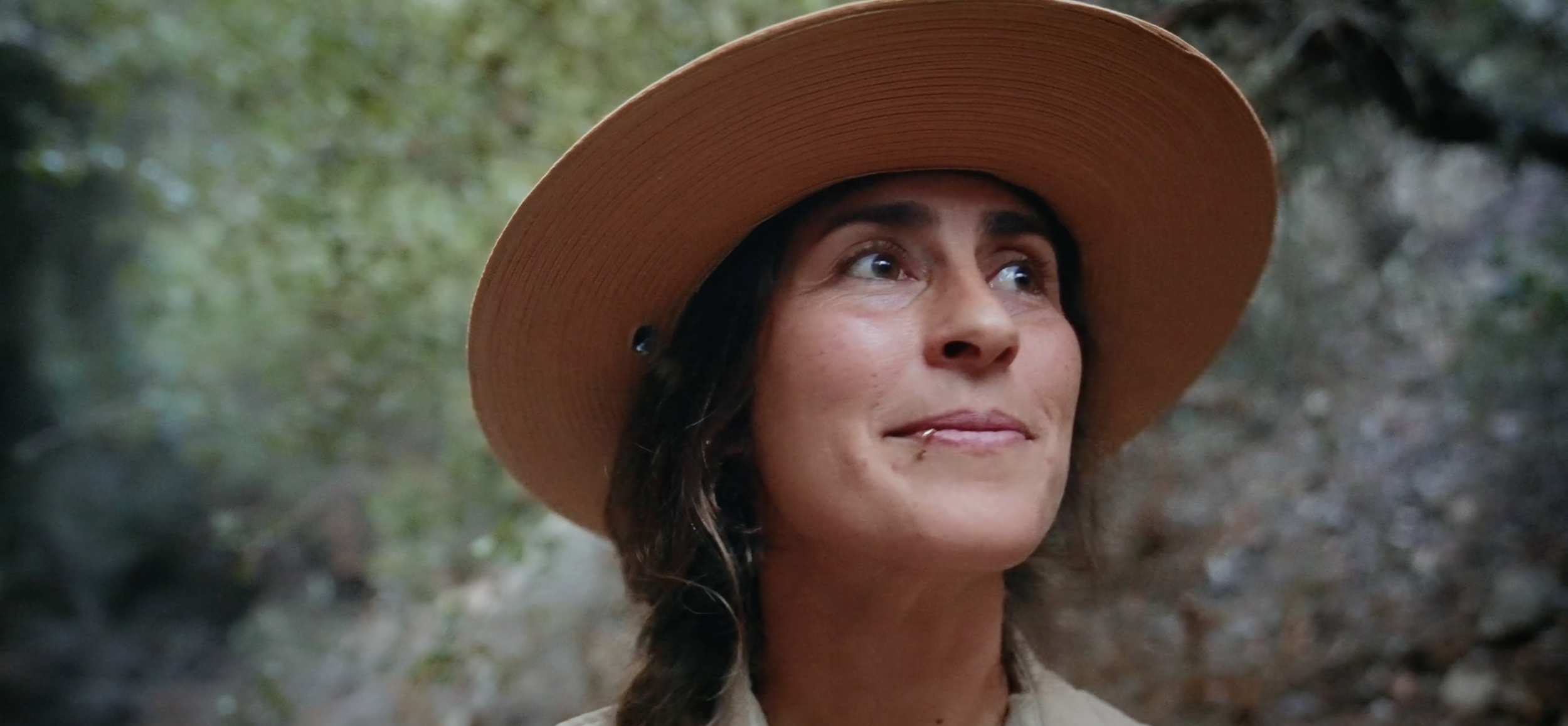 A woman wearing a wide-brimmed straw hat outdoors with greenery in the background.