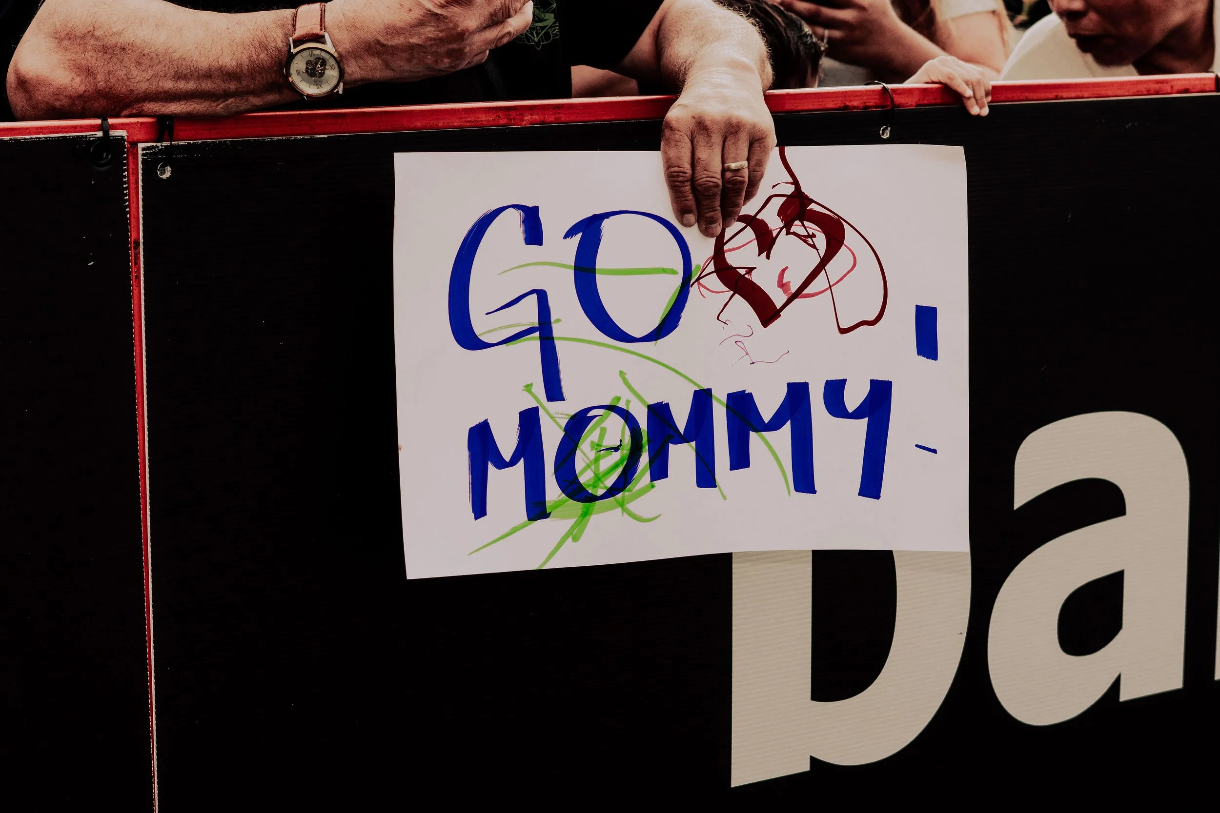 A close-up of a person's hand holding a sign with colorful writing that says 'G0AL' with a drawing of a face and green lines, in front of a scoreboard with a black background.