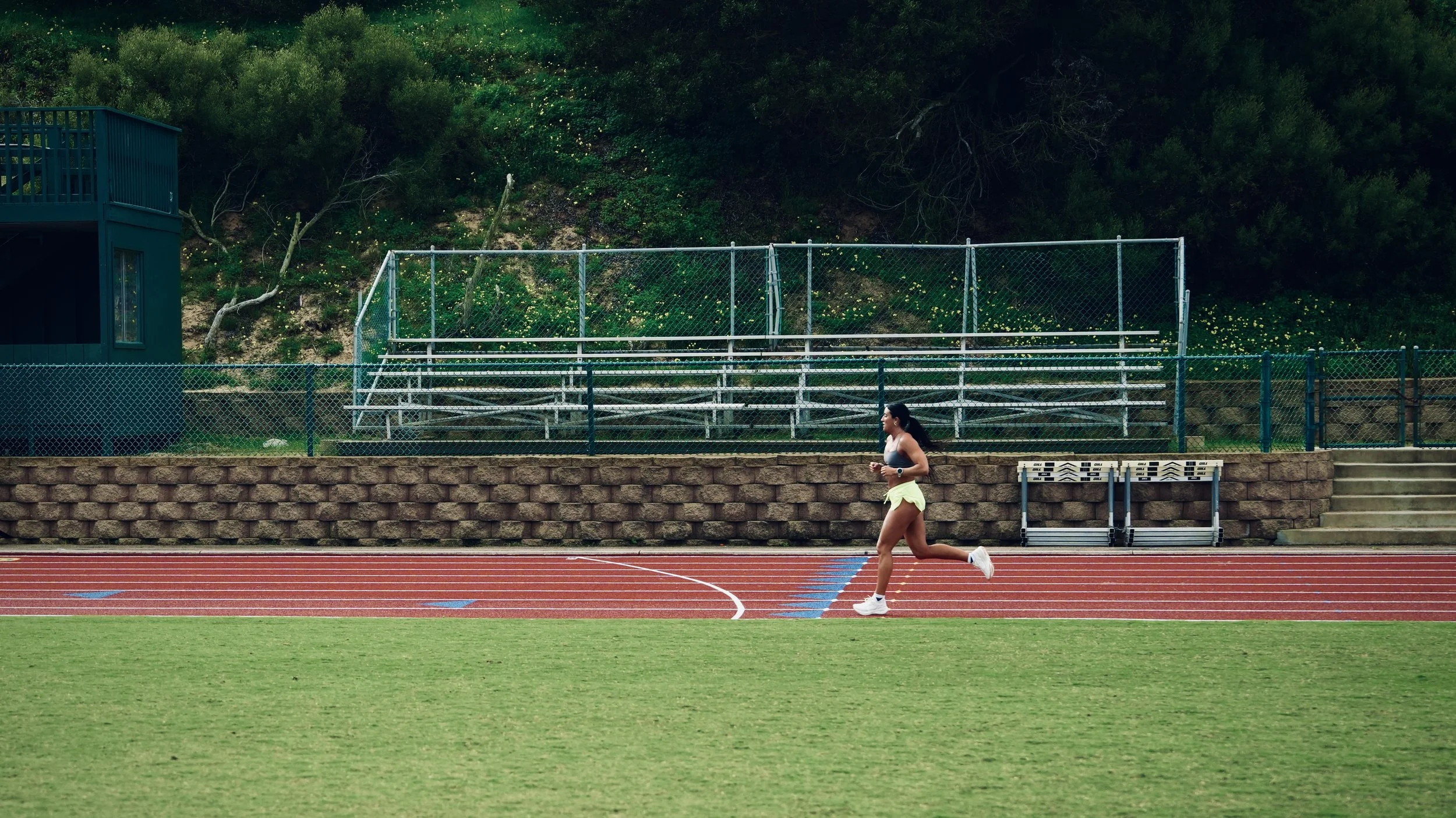A woman in athletic clothing running on a red track at a sports stadium with green grass and bleachers in the background.