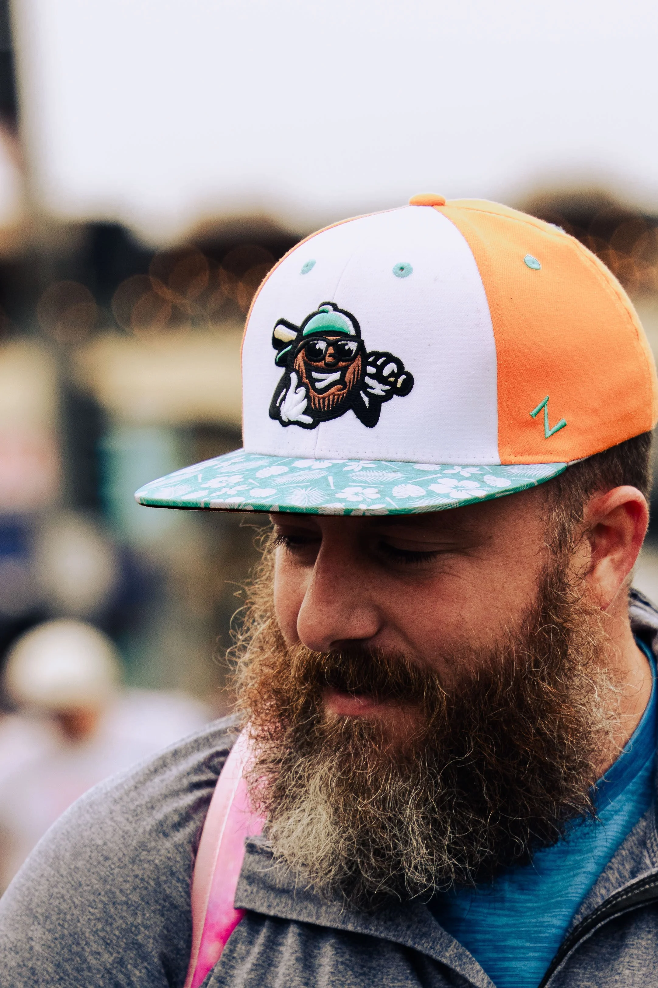 A man wearing a grey hoodie, a pink backpack, and a baseball cap with a cartoon pirate on the front, with a blurred background of lights.