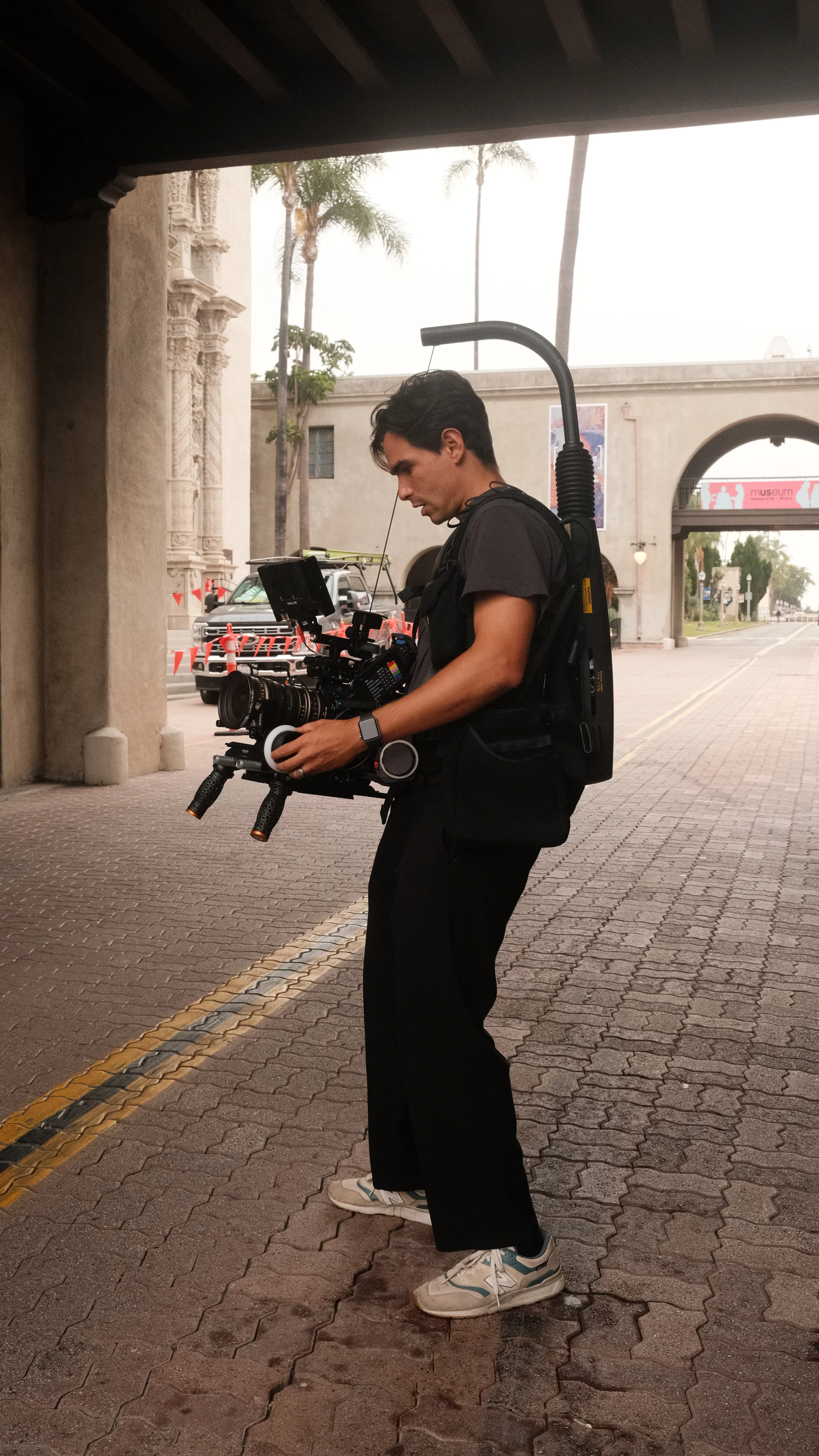 A young man holding a professional film camera, standing outdoors on a brick sidewalk with palm trees and a building in the background.