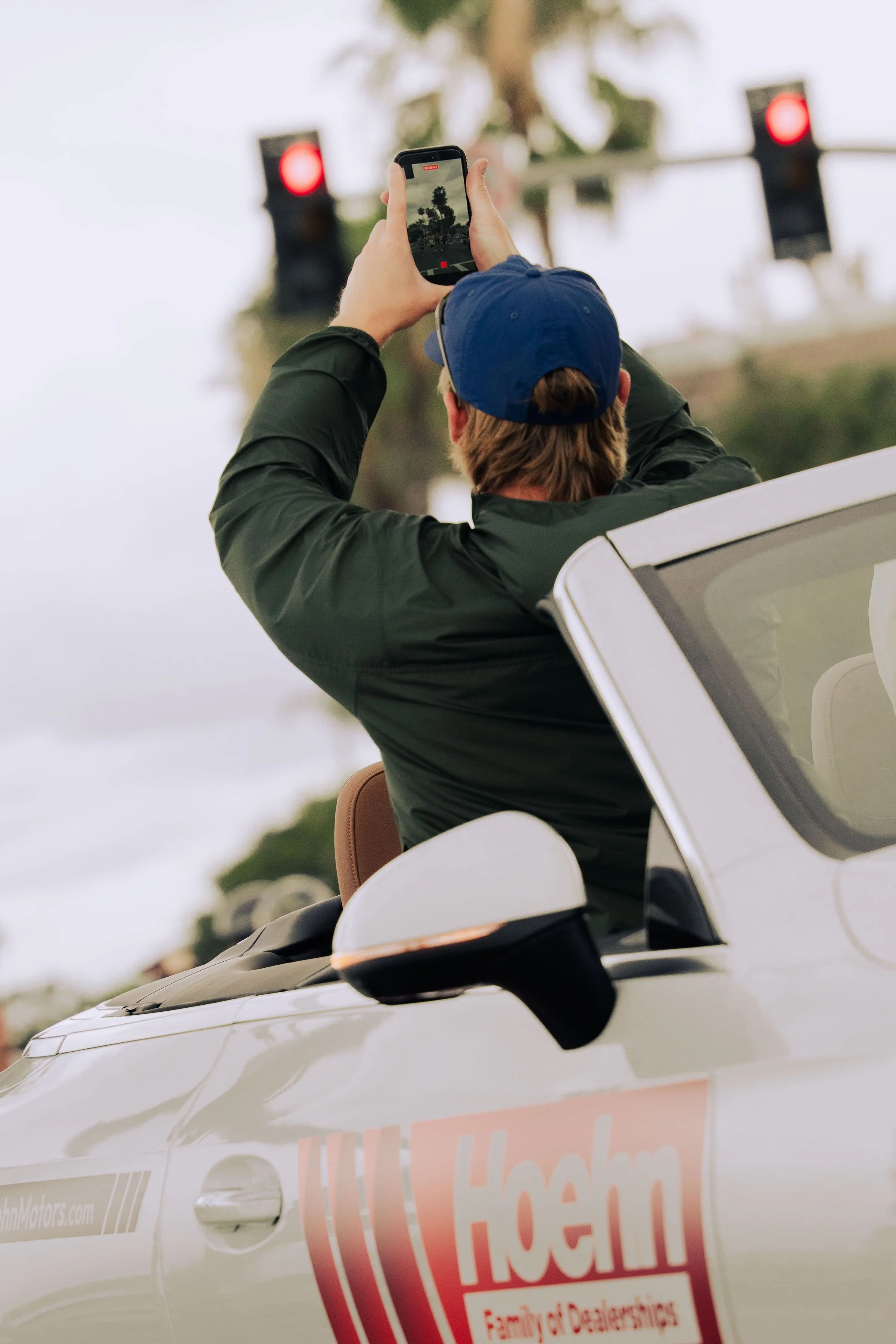 A person wearing a blue cap and dark jacket is taking a photo with a smartphone while sitting in a white vehicle, with a blurred background of trees and a red traffic light.