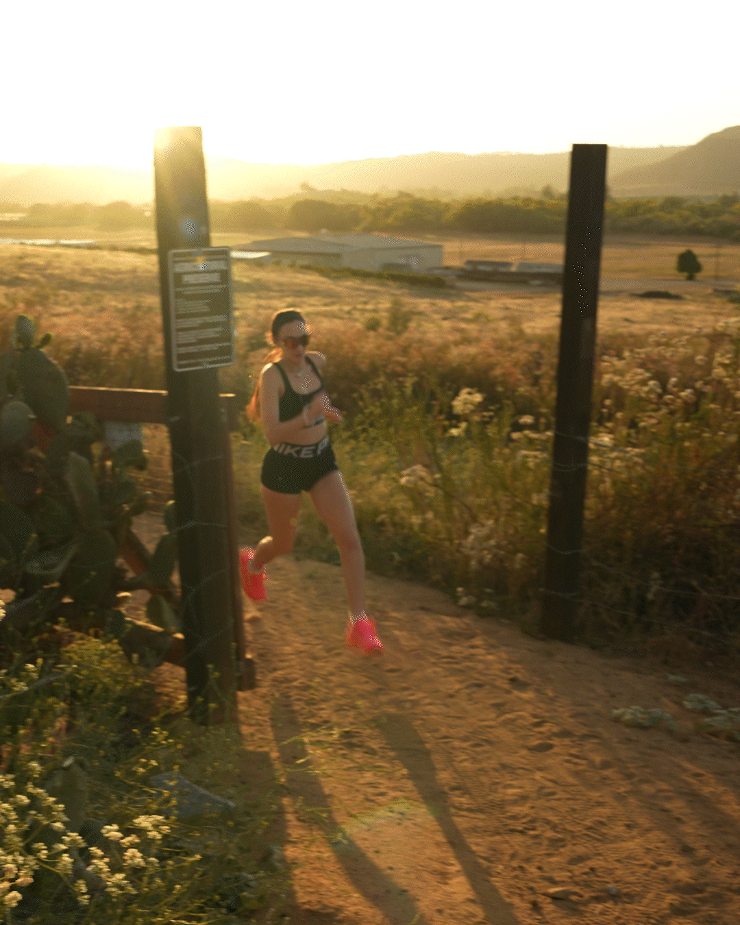 A woman running along a dirt trail during sunset, wearing sunglasses, a black sports bra, black shorts with white lettering, and bright pink running shoes.