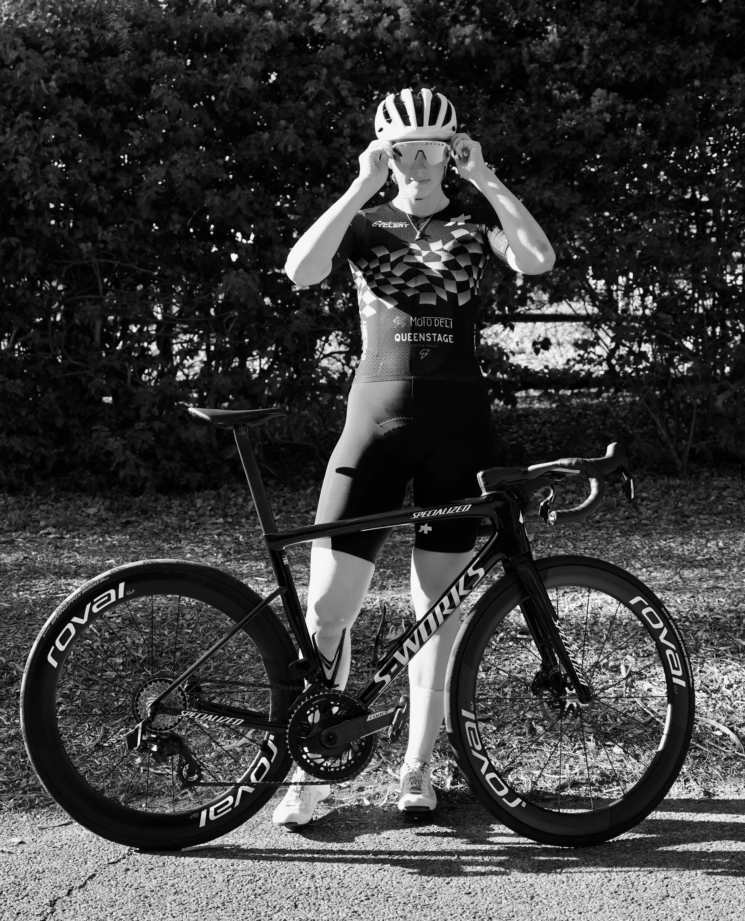 A cyclist in cycling apparel and helmet adjusting sunglasses while standing with a bicycle on a paved path in front of dense foliage.