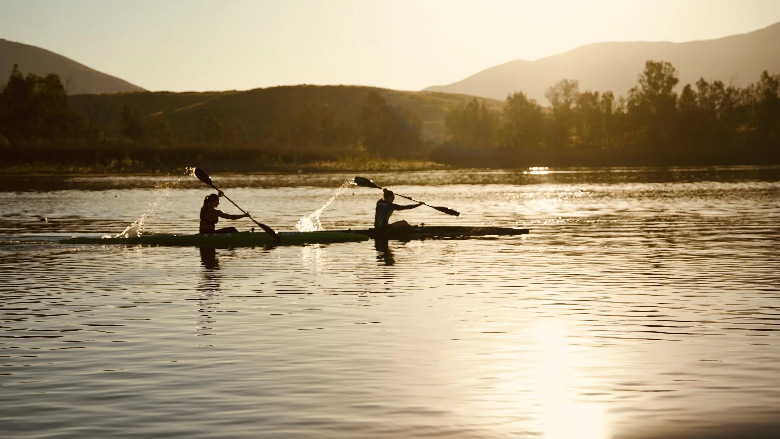 Two people kayaking on a calm lake during sunset with mountains and trees in the background.