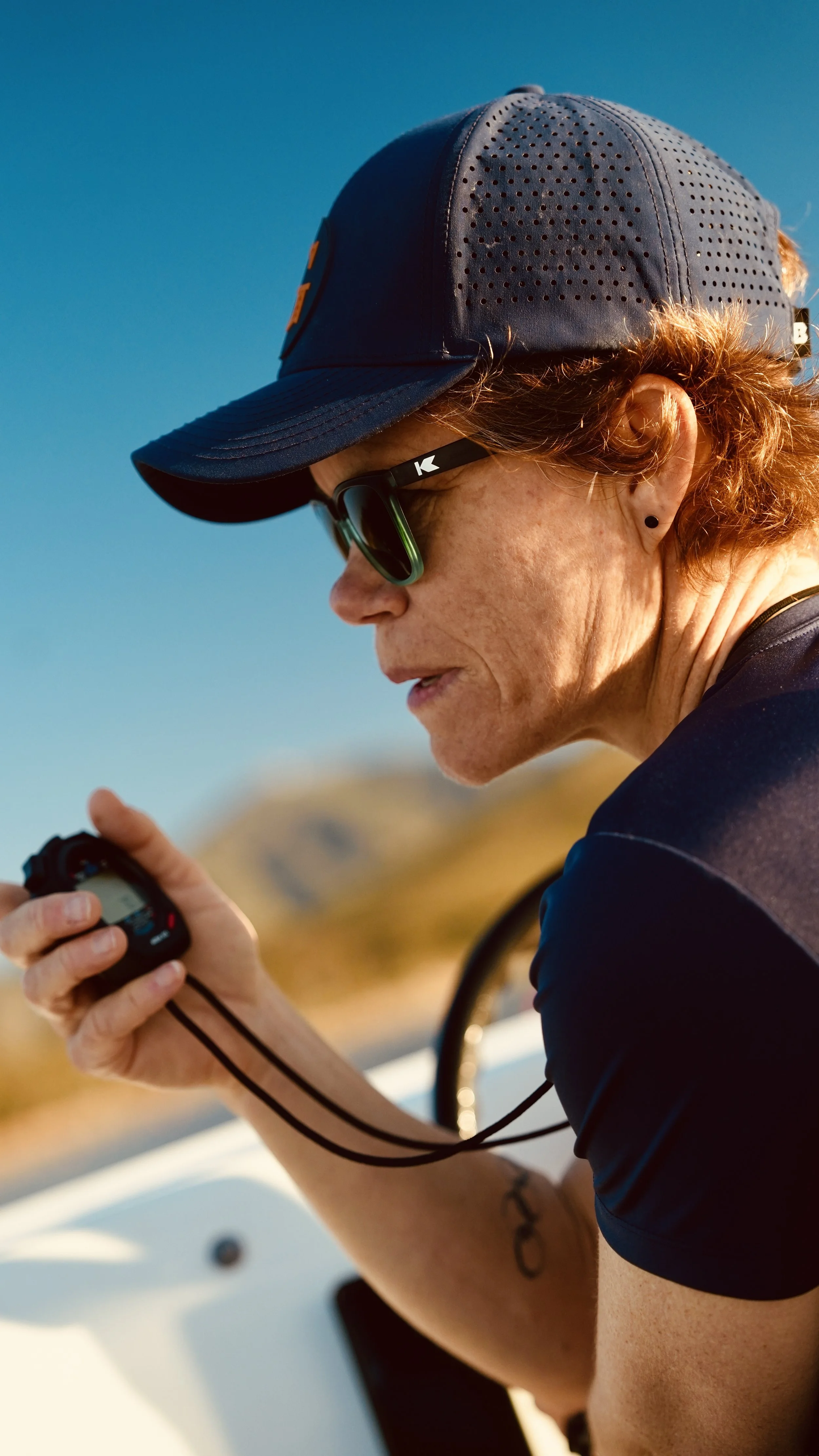 A woman with curly red hair, wearing sunglasses, a blue cap, and a black shirt, is looking at a device in her hand, likely a music player or phone, outdoors with a blue sky and blurred landscape in the background.