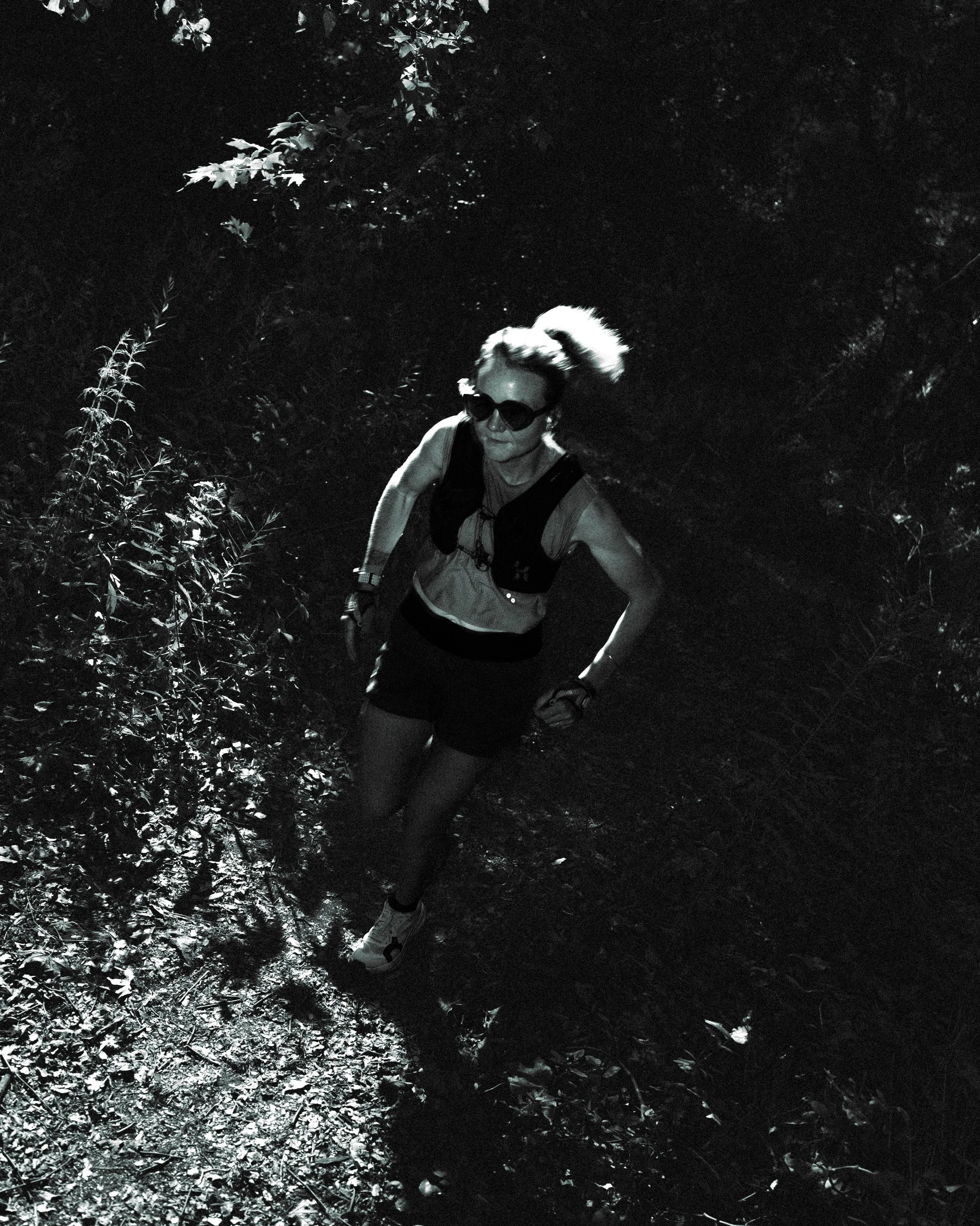 A woman hiking at night in a wooded area, wearing sunglasses, a vest, and shorts, illuminated by a light source.