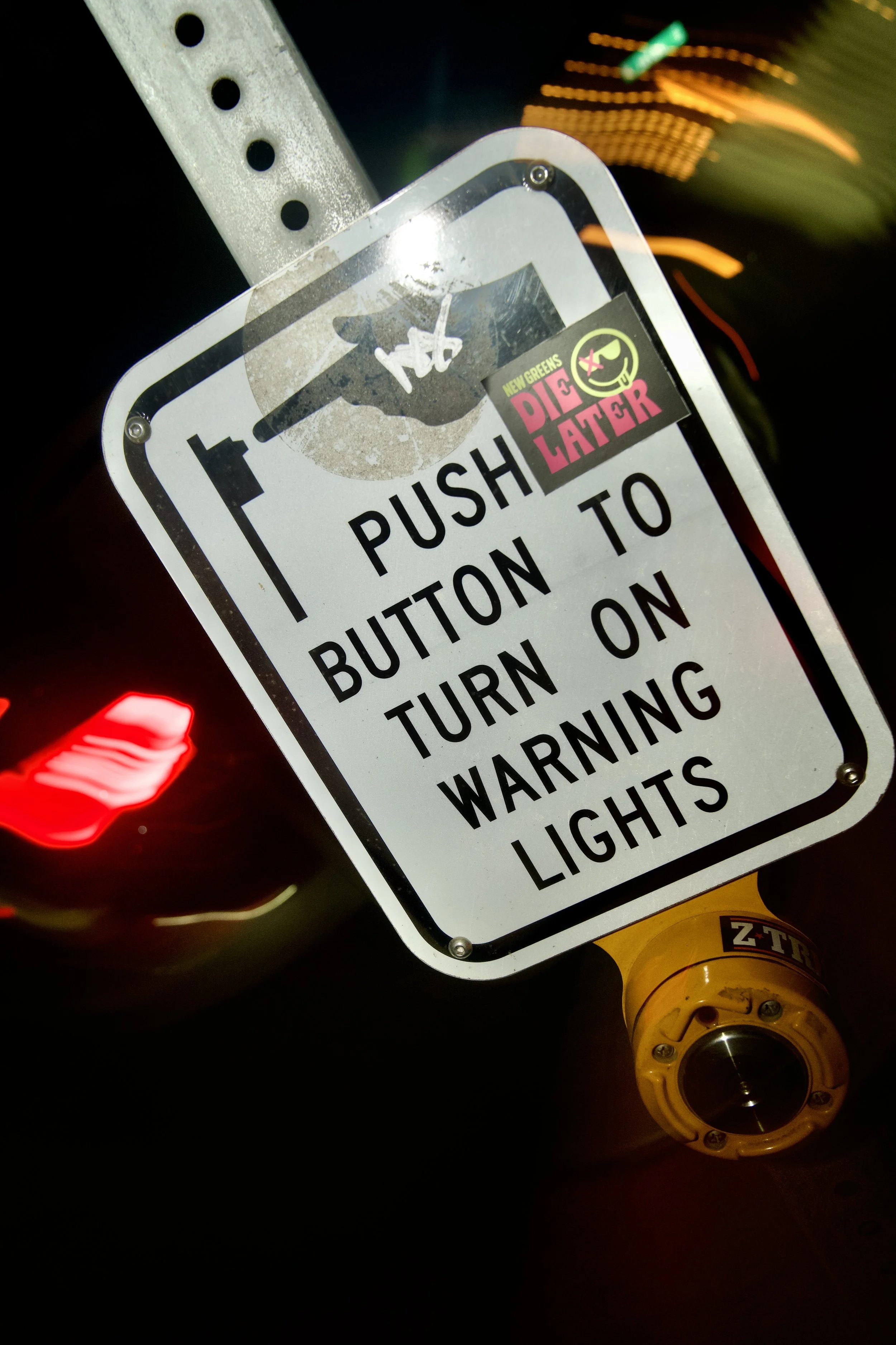 Close-up of a street sign with a sticker that reads "Die Later" with a smiley face and the words "New Greens". The sign instructs to push a button and turn on warning lights, with blurry background lights.