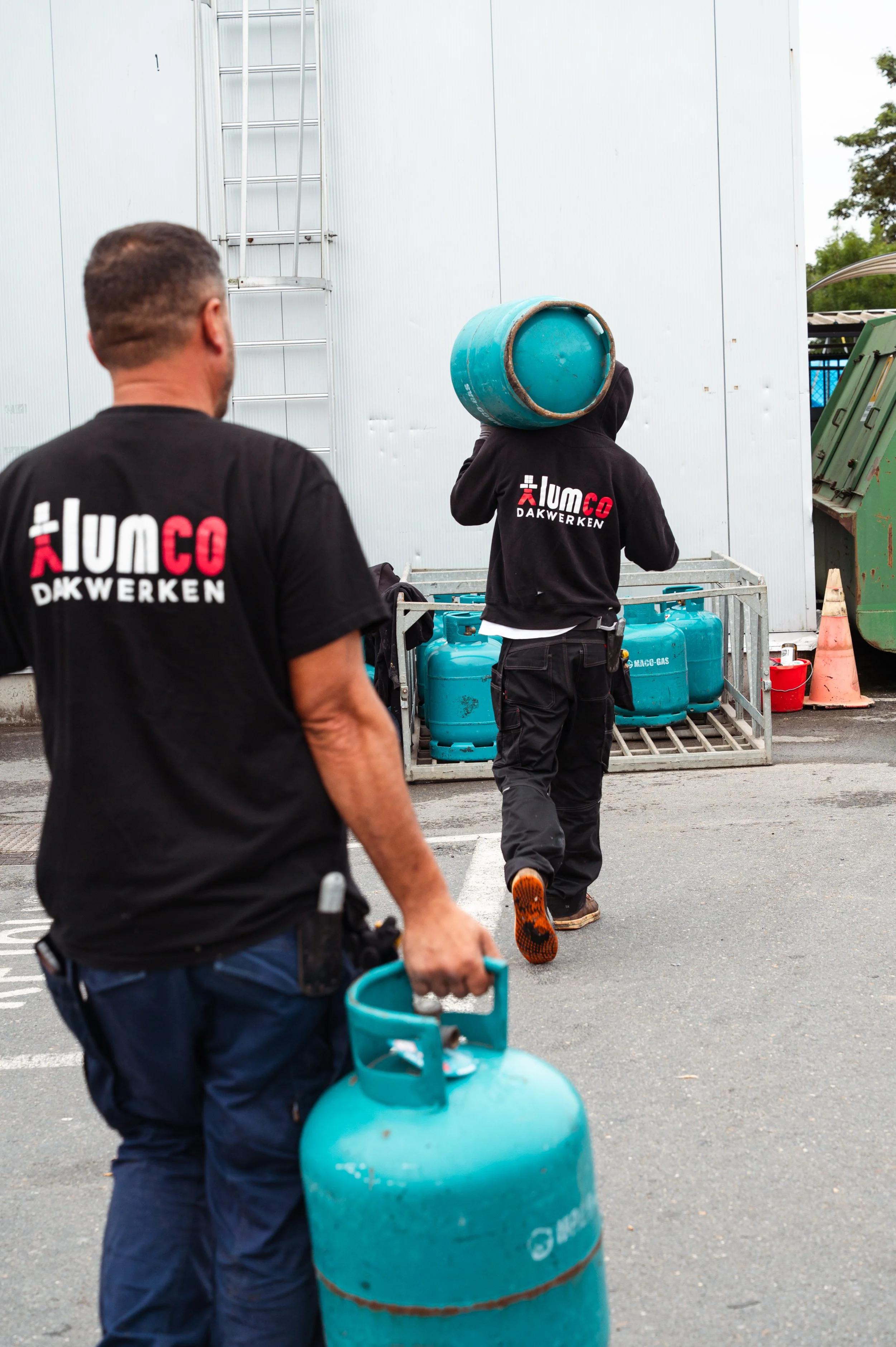 Twee mannen met T-shirts met het logo 'lumco DAKWERKEN' op het werk. Eén man draagt een grote blauwe gasfles op zijn schouder, de ander houdt een vergelijkbare flessen vast.