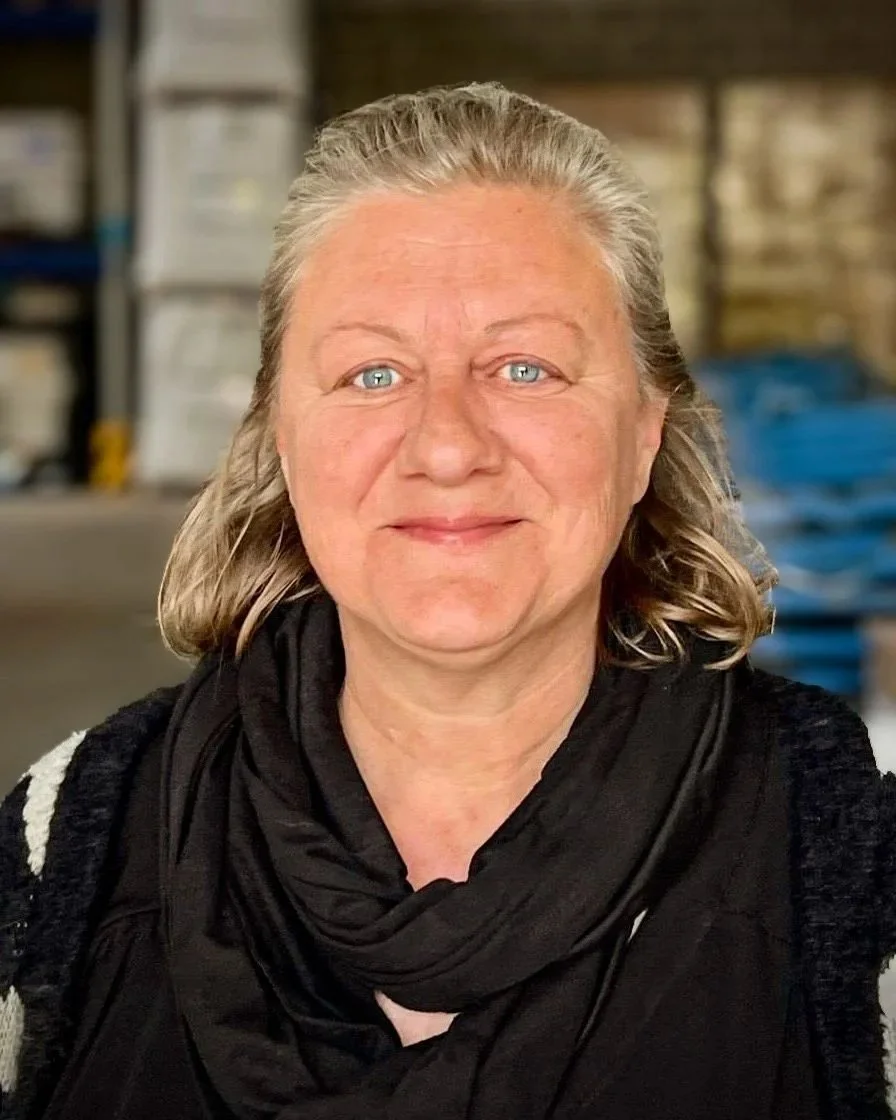Vrouw met rood haar en witte blouse, glimlachend met armen gekruist, binnen een houtzagerij of meubelmakerij.